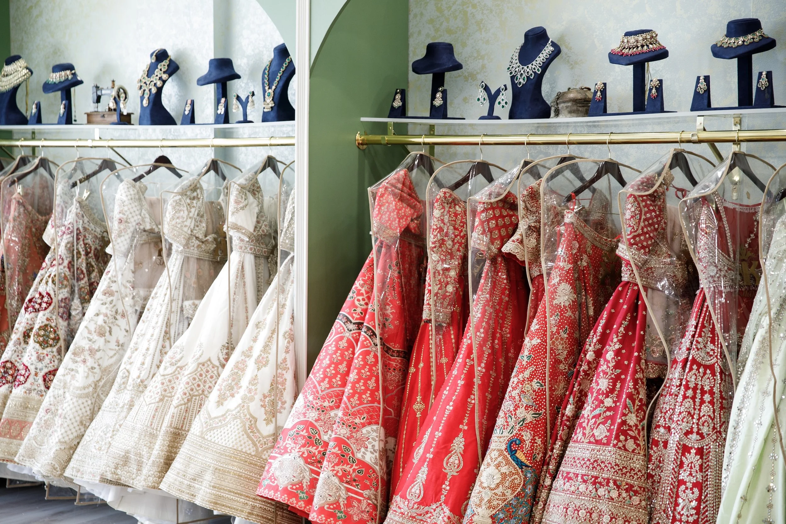 A display of traditional Indian wedding dresses and jewelry in a boutique with gowns hanging on a rack and ornate necklaces on mannequin busts.