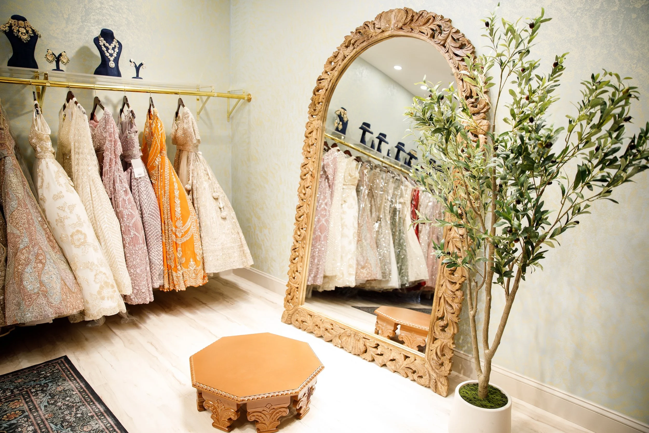 Luxury boutique fitting room with ornate mirror, display of elegant dresses on racks, decorative jewelry mannequins, green potted plant, small wooden stool, and a patterned rug.
