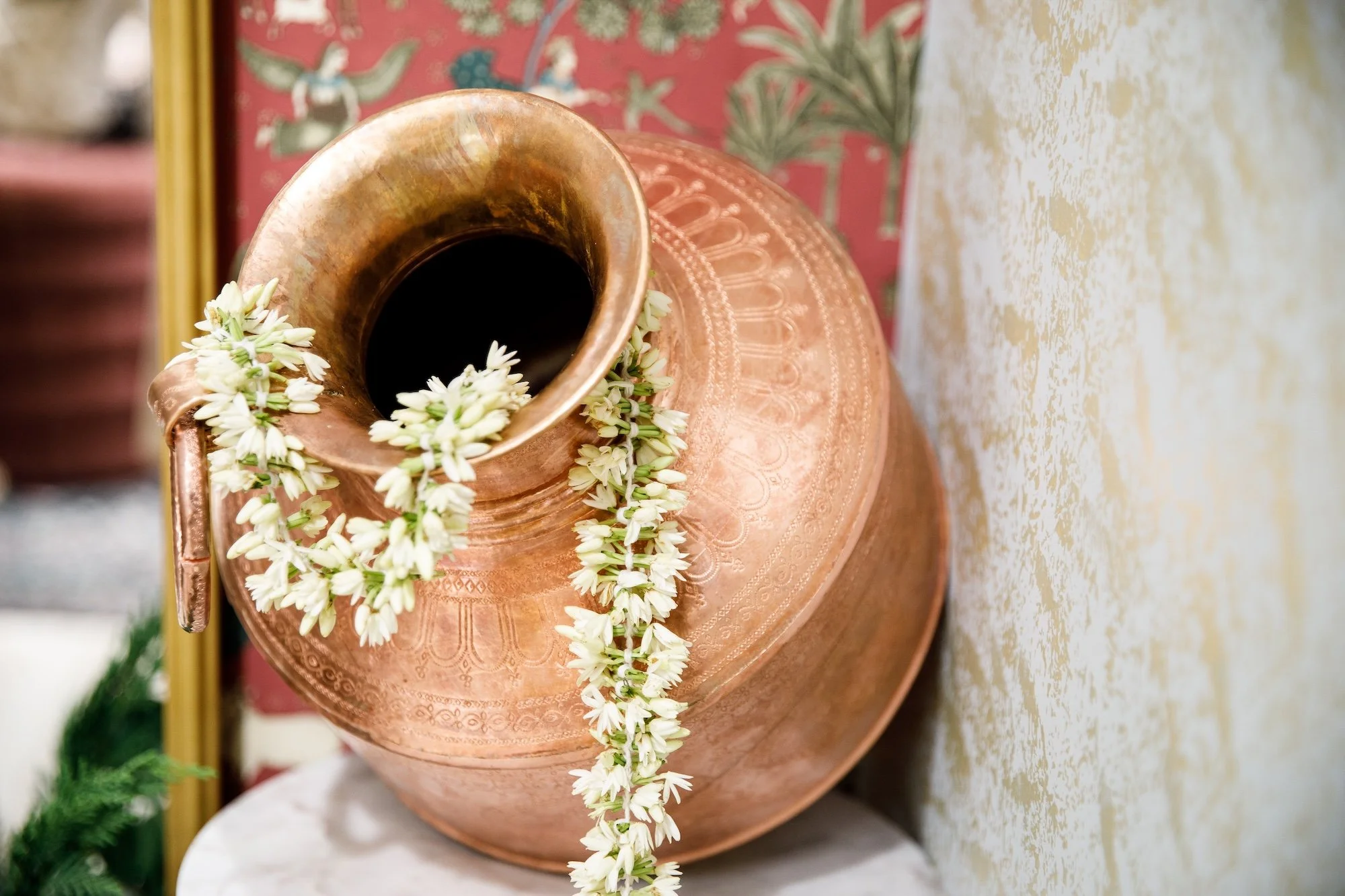 A copper water jug decorated with a strand of white flowers, placed on a white round surface, with a patterned red cloth and beige textured wall in the background.