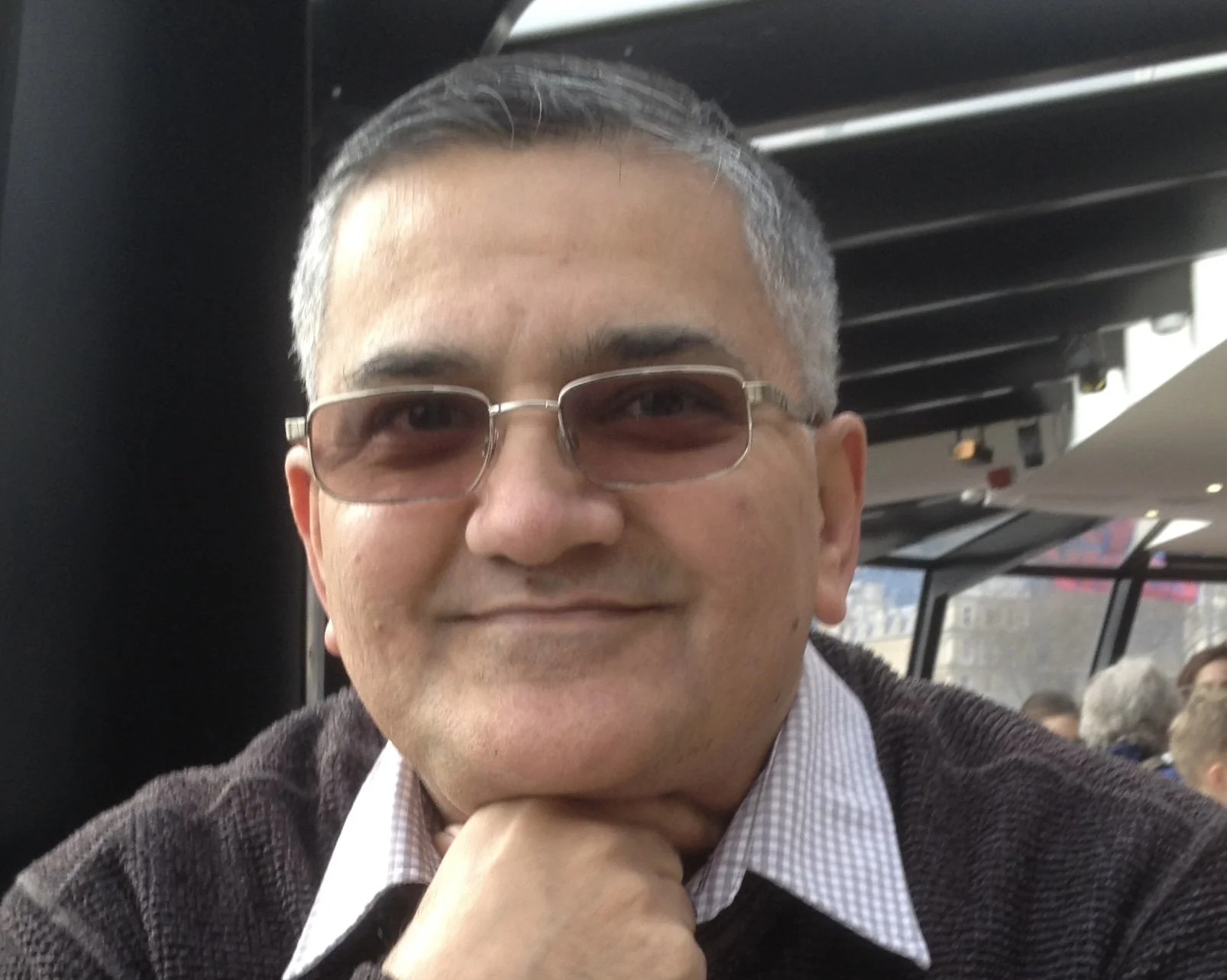 A man smiling and wearing glasses, sitting indoors in a public space with other people and large windows in the background.