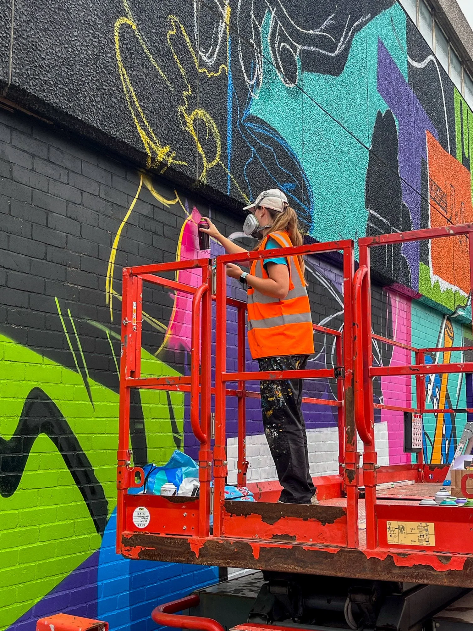 Brittany from K&B Design wearing a safety vest and a respirator mask is standing on a red lift, spray painting a colourful mural on a brick wall in Ipswich for the wild walls mural festival.