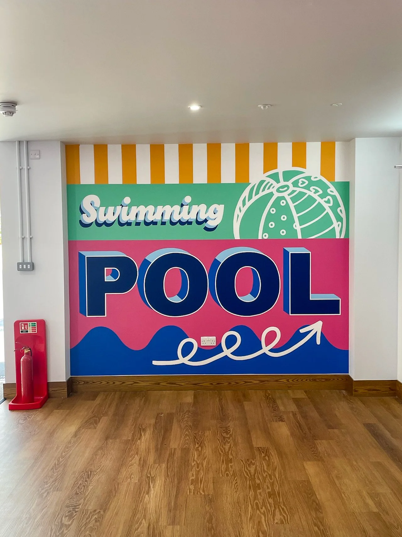Way finding signage / wall mural for Haven, Kent Coast, with the words 'Swimming Pool' and a drawing of a beach ball, vibrant pink, green, blue, and yellow design.