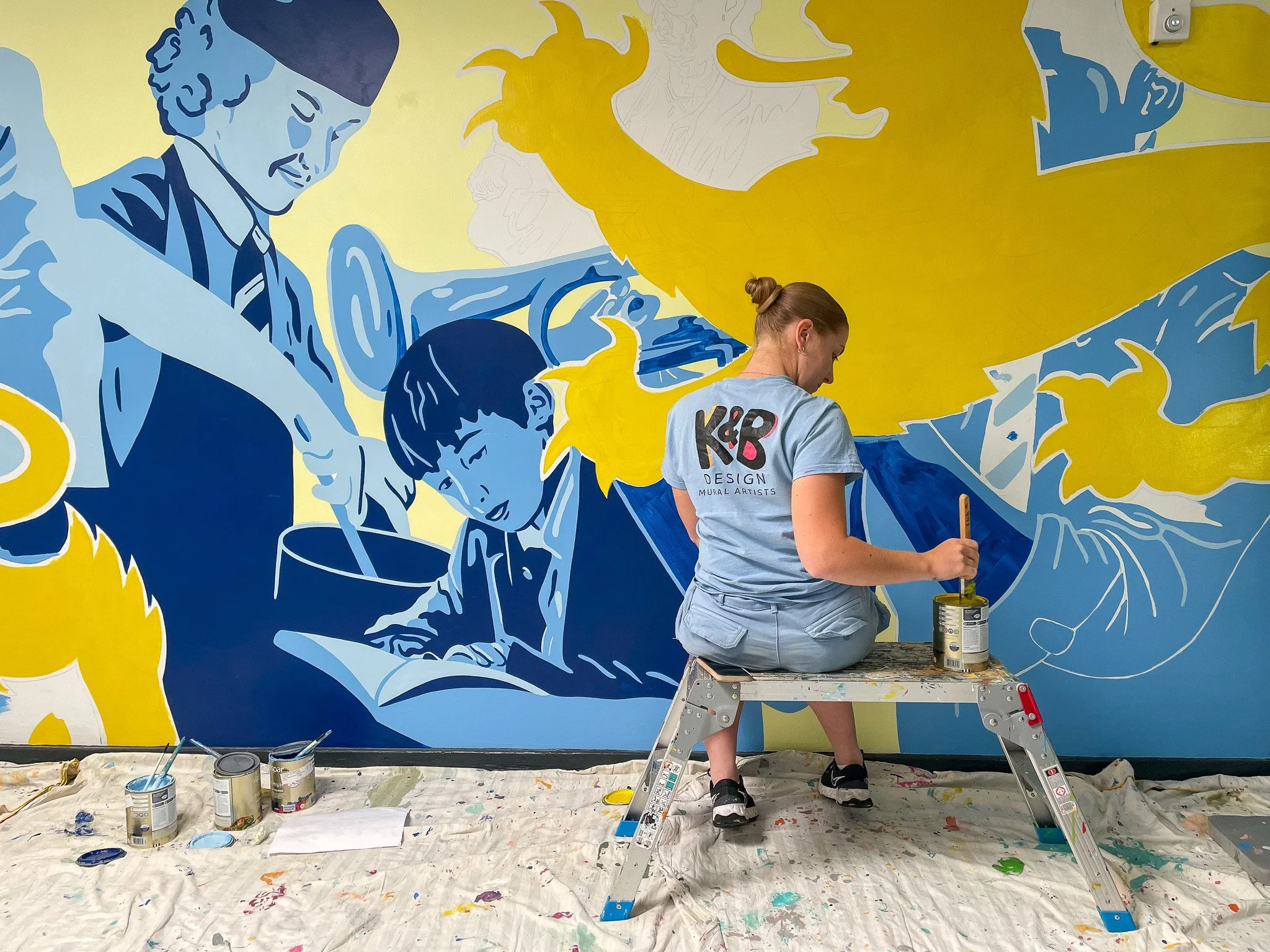 Kirsty from K&B Design, painting a large mural on a school wall. The mural depicts stylised blue and yellow illustrations of people, including a woman pouring a drink and a boy reading a book.