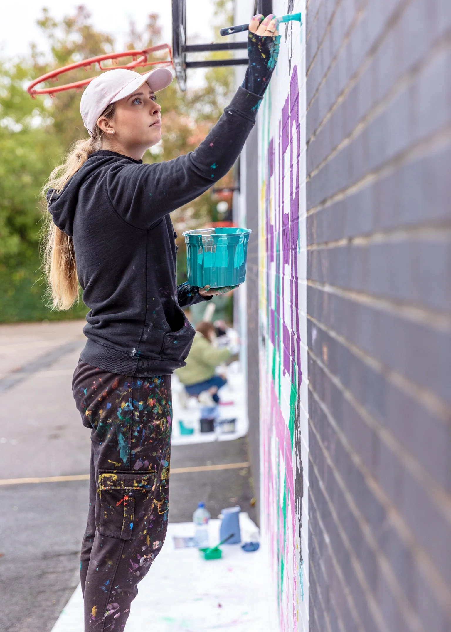 Brittany from K&B Design painting a colourful playground mural on a brick wall outdoors, wearing a white cap, black hoodie, and paint-spattered pants, holding a paintbrush and a bucket of teal paint.