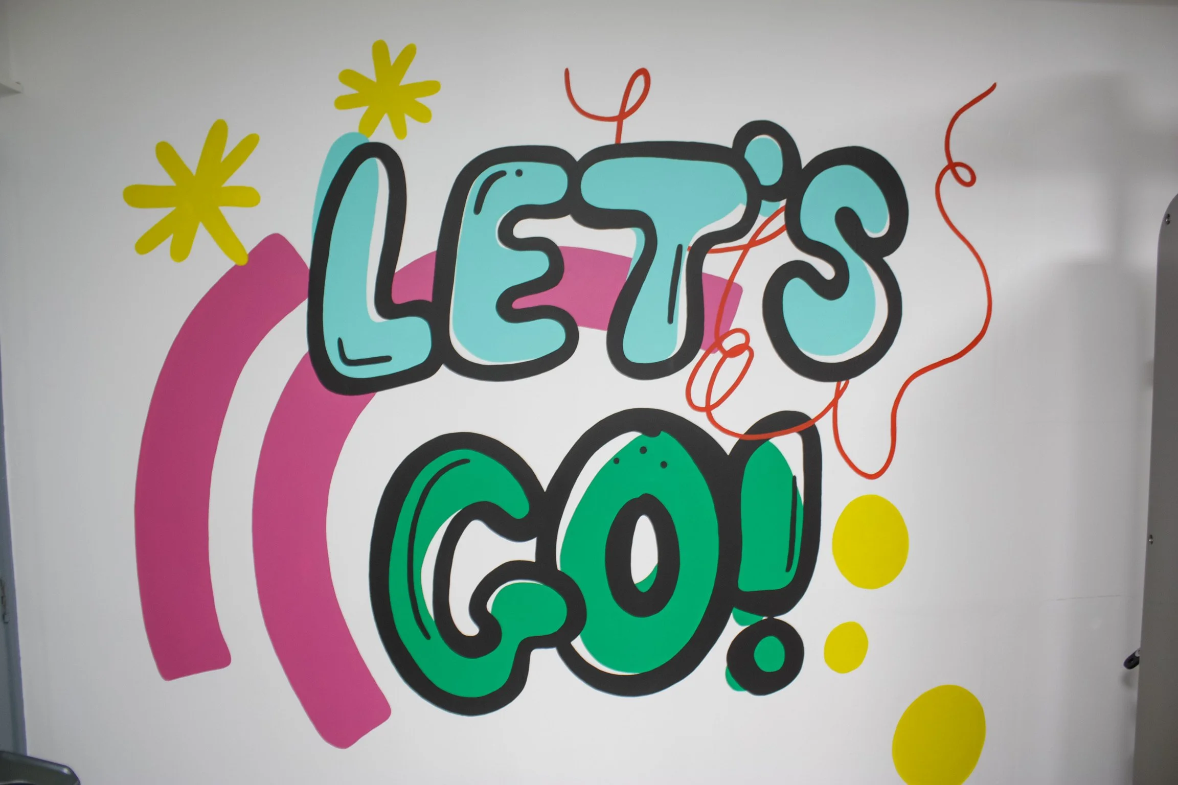 Colourful graffiti-style wall art mural with the words "LET'S GO!" in bold letters, featuring pink, green, blue, yellow, and red accents and decorative elements.