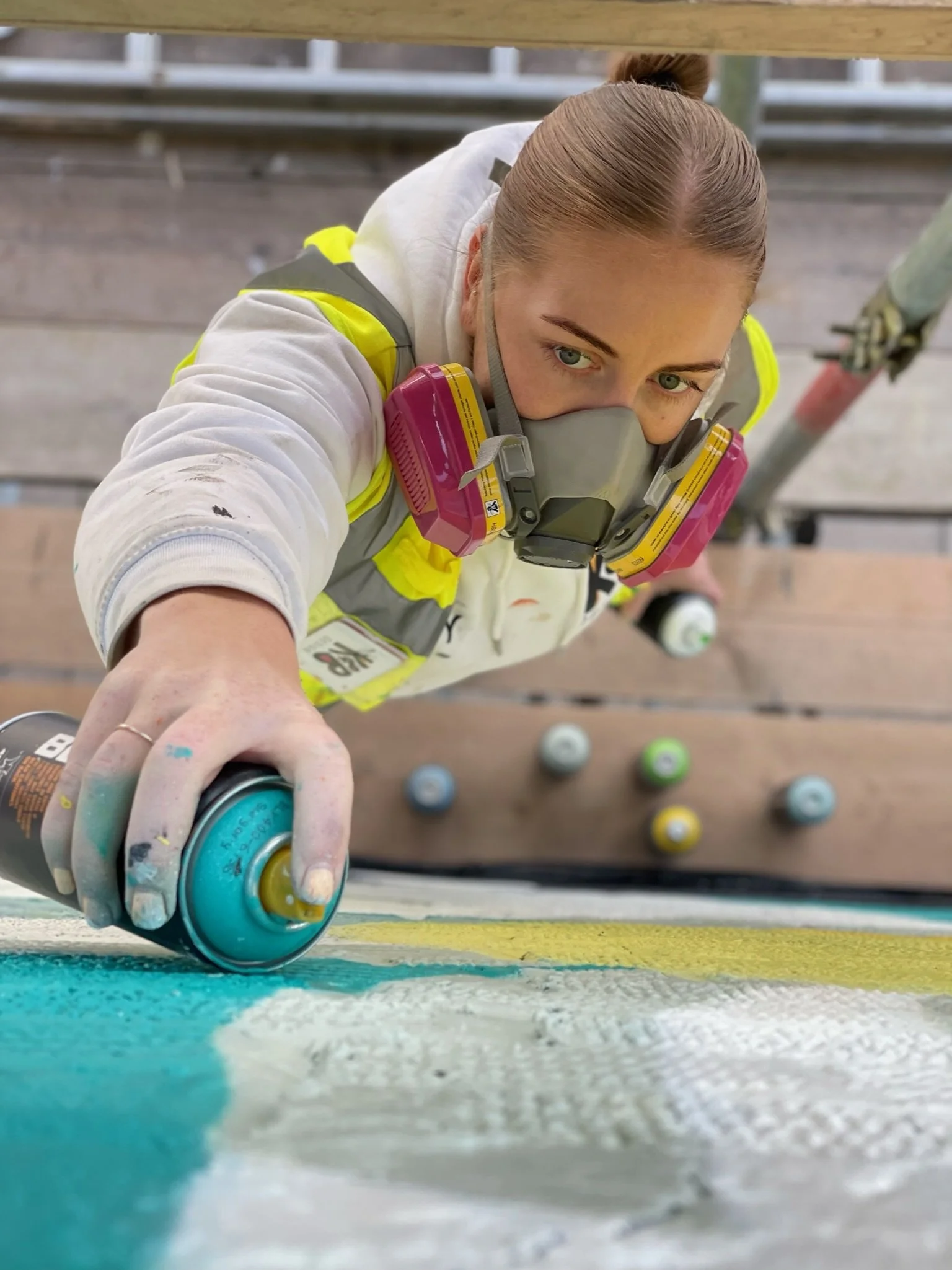 Kirsty from K&B Design wearing a respirator mask and safety vest spray painting a surface with turquoise paint. She is looking up from a scaffold, with spray paint cans on the ground below.