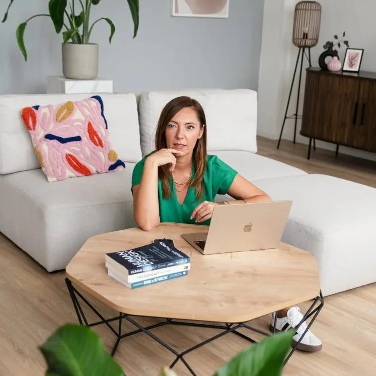 A woman in a green shirt sitting at a wooden coffee table with a laptop and books, in a cozy living room with a white sofa, colorful decorative pillow, plant, and modern furniture.