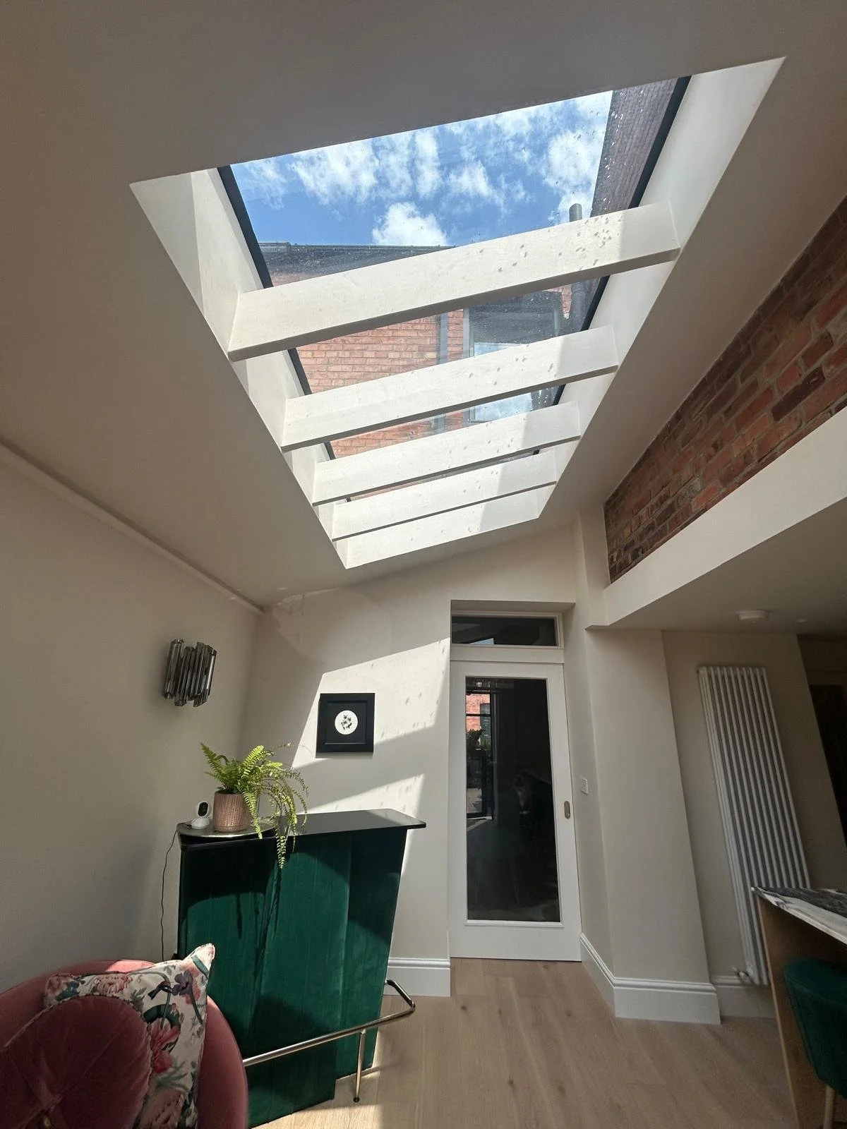 Interior of a room with a large skylight window in the ceiling, revealing a view of the blue sky with clouds and neighboring brick buildings. The room has white walls, wooden flooring, and some furniture including a green desk or cabinet with a potte