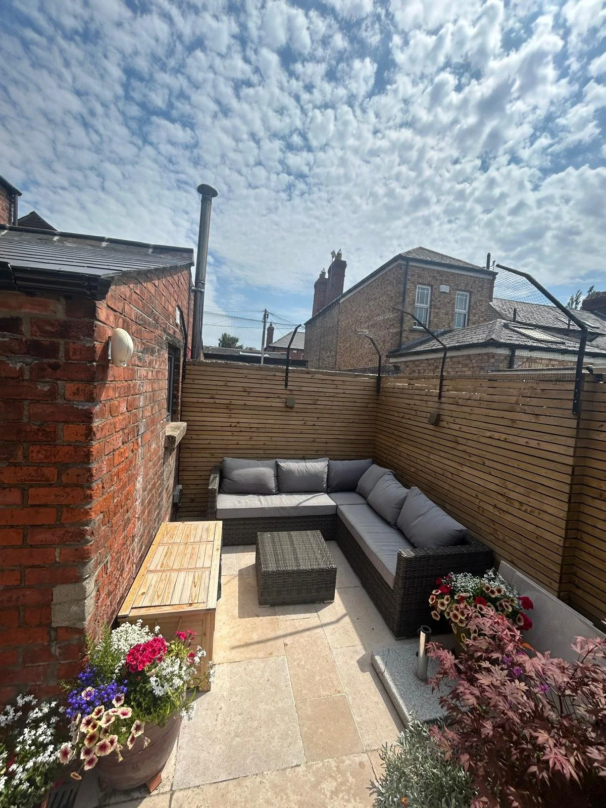 A small outdoor patio with a gray sectional sofa, a wicker table, and potted colorful flowers, enclosed by wooden fencing and brick walls, with a partly cloudy sky overhead.