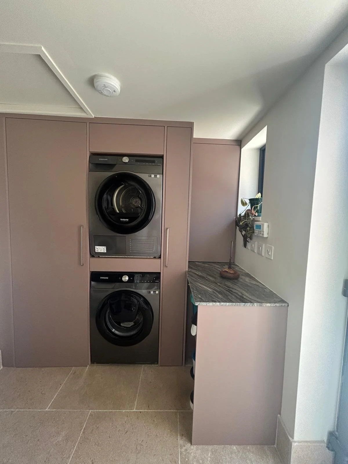 A laundry room with two stacked Samsung front-loading washers and dryers, beige cabinetry, a white wall with a small window, and a countertop with a plant and a wall-mounted thermostat.