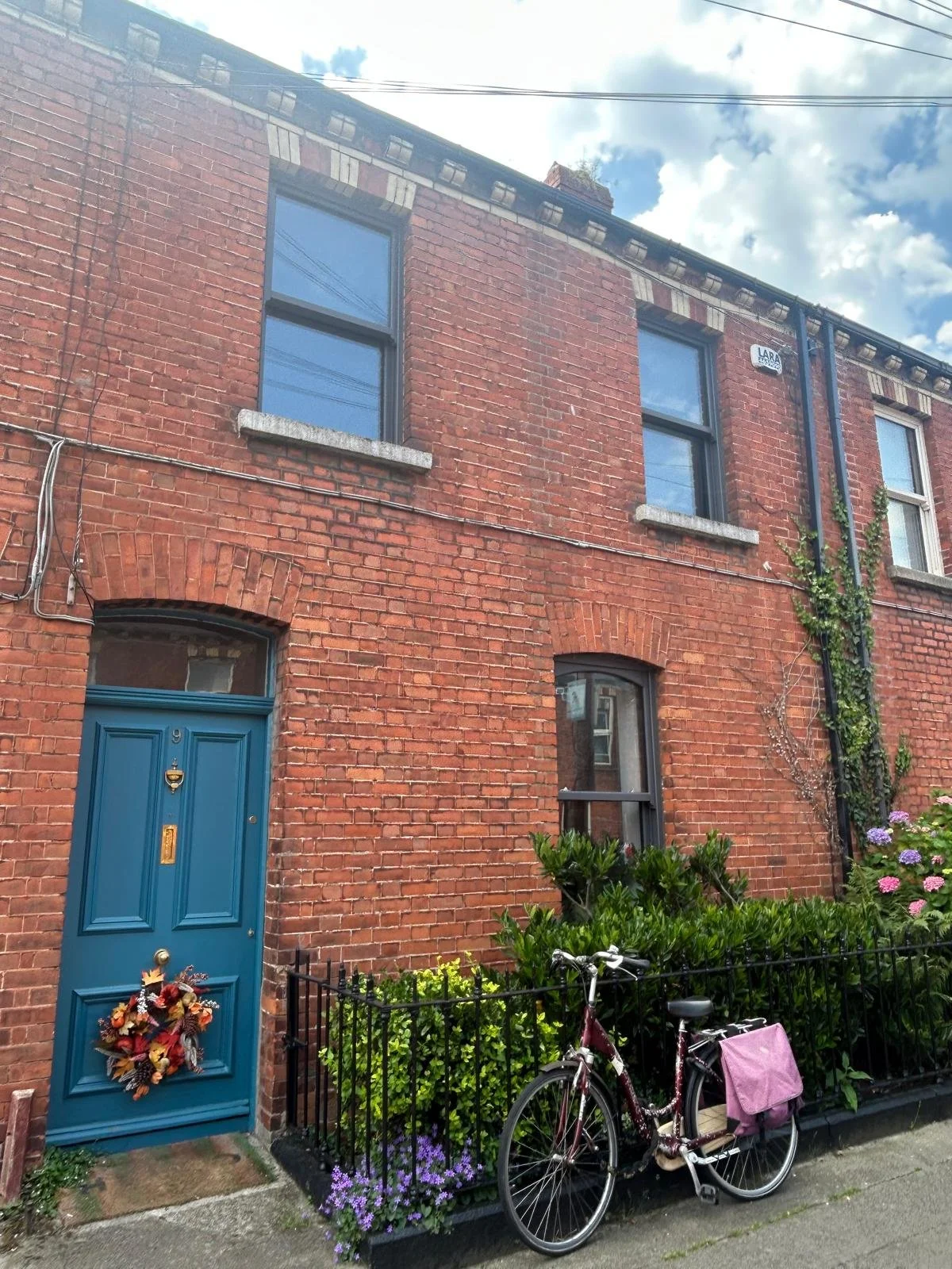 A red brick multi-story building with blue doors and window frames, a black fence with purple flowers, a bicycle, and green plants outside, under a partly cloudy sky.