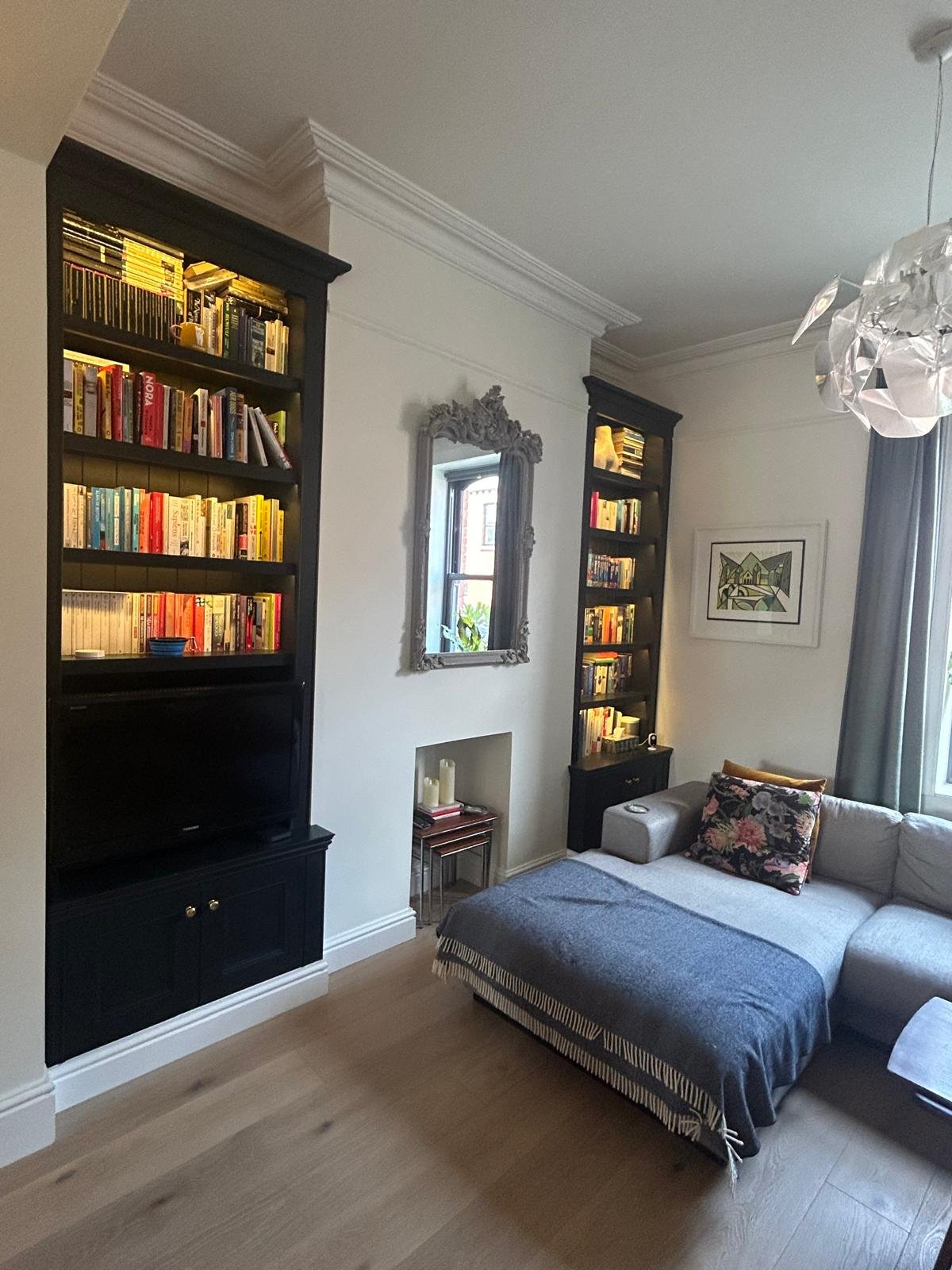 Living room with a black bookshelf filled with books, a decorative mirror, a small side table with candles, a framed artwork, a large window with curtains, a gray sofa with floral cushions, and a modern chandelier.
