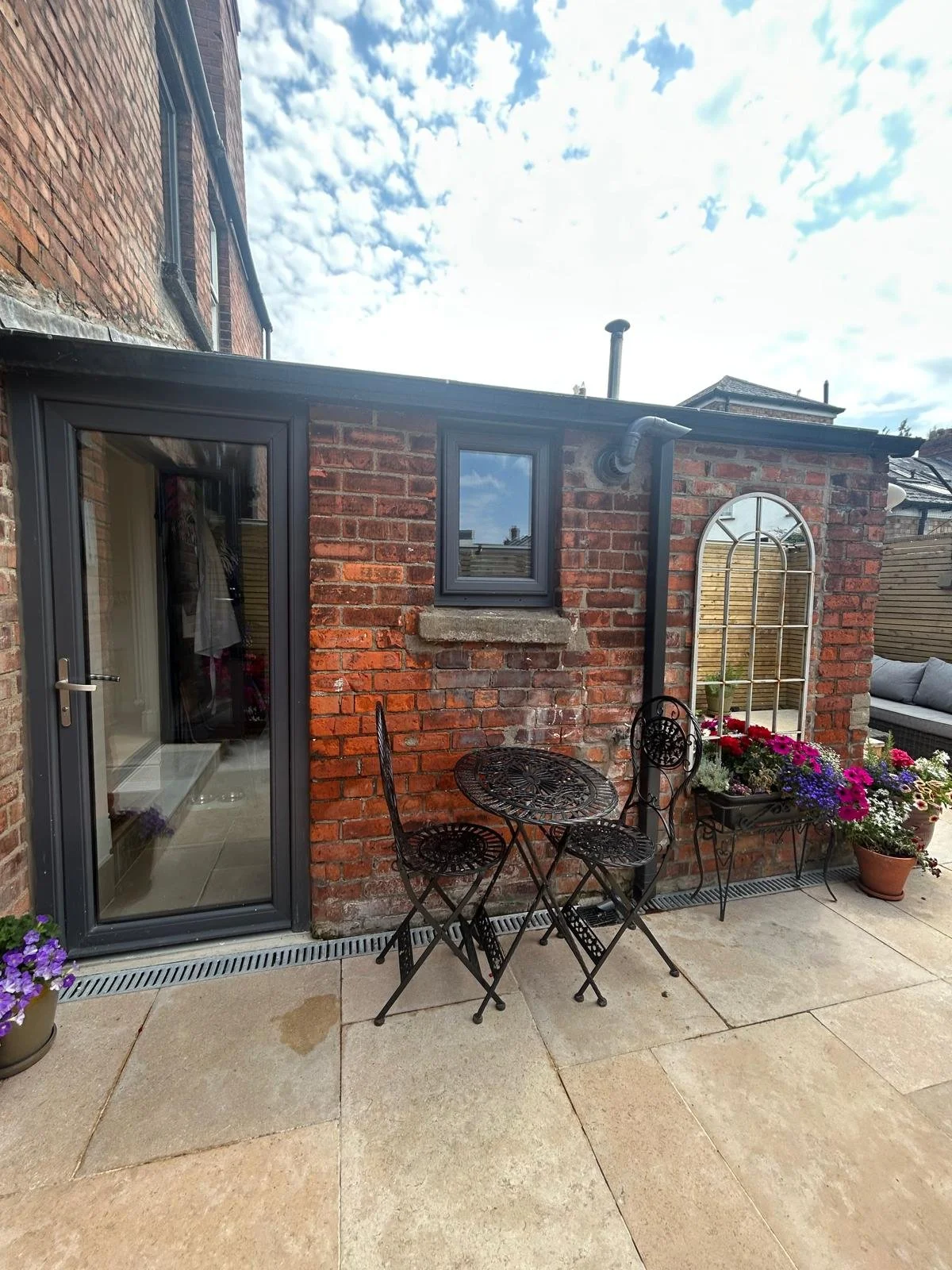 A small outdoor patio area with a black metal table and two chairs, decorated with colorful potted flowers, next to a brick house with a glass door and window, under a partly cloudy sky.