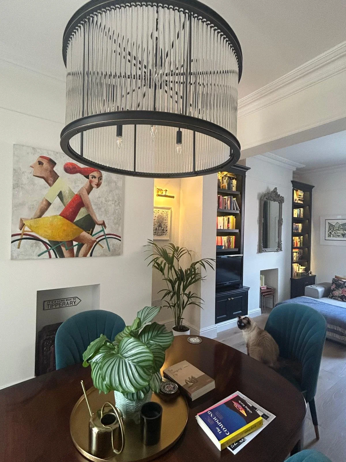 Living room with a large chandelier, a colorful modern painting of people riding bicycles on the wall, a black bookshelf, a mirror, a beige sofa, a cat sitting on a blue chair, a round wooden table with a green potted plant and books, and various dec