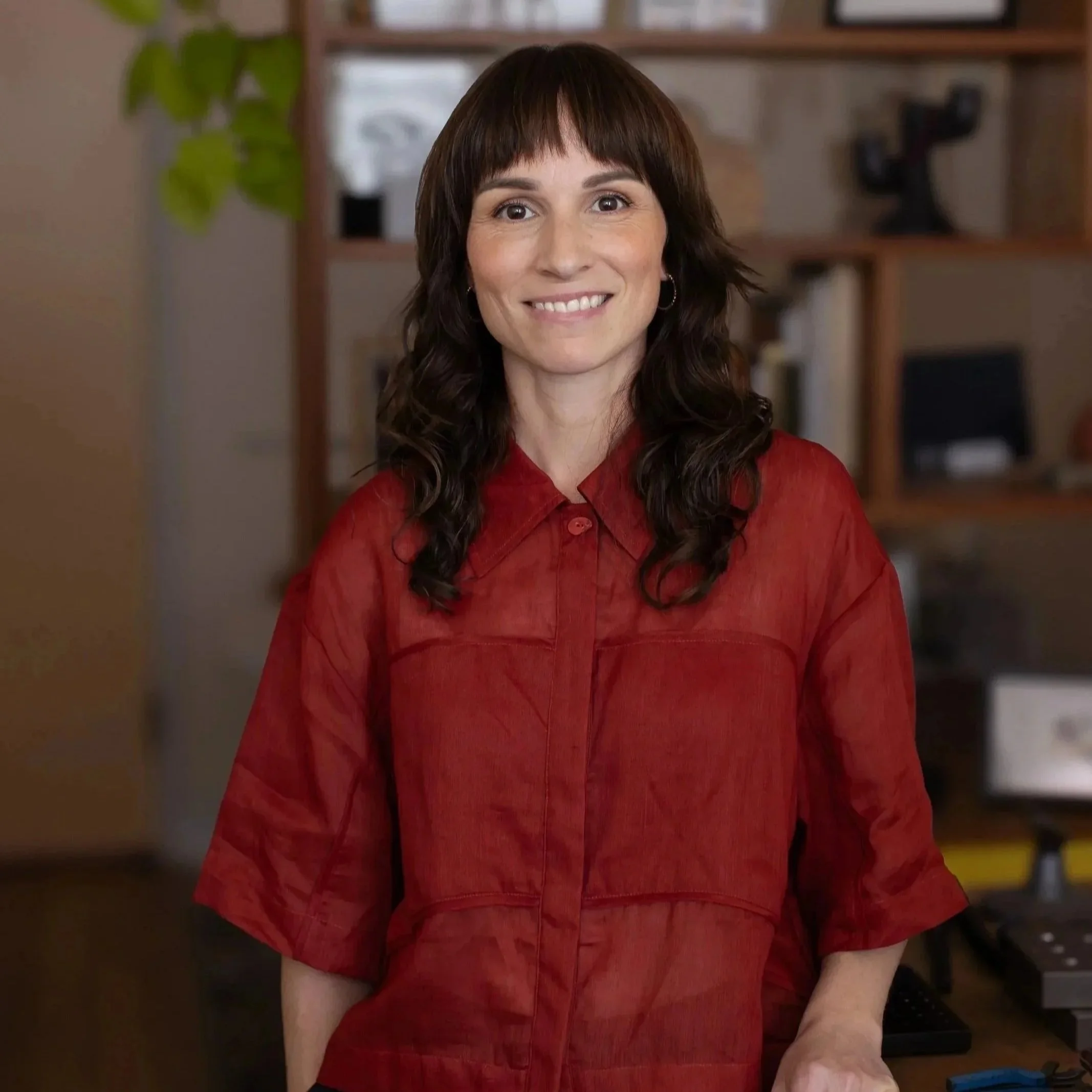 A woman (Kate, the Jeweller) with shoulder-length dark hair and bangs, wearing a red shirt, smiling in a cozy indoor jewellery studio setting with shelves and a plant in the background.