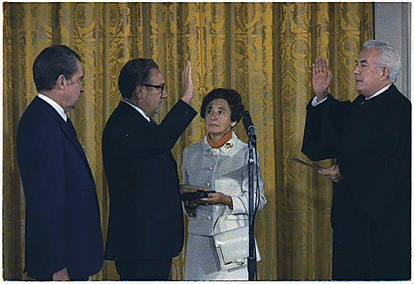 Henry Kissinger is sworn in as Secretary of State under Richard Nixon, who is by his side. Kissinger is raising his right hand, his mother is holding a bible, and the Supreme Court Chief Justice is delivering the oath.