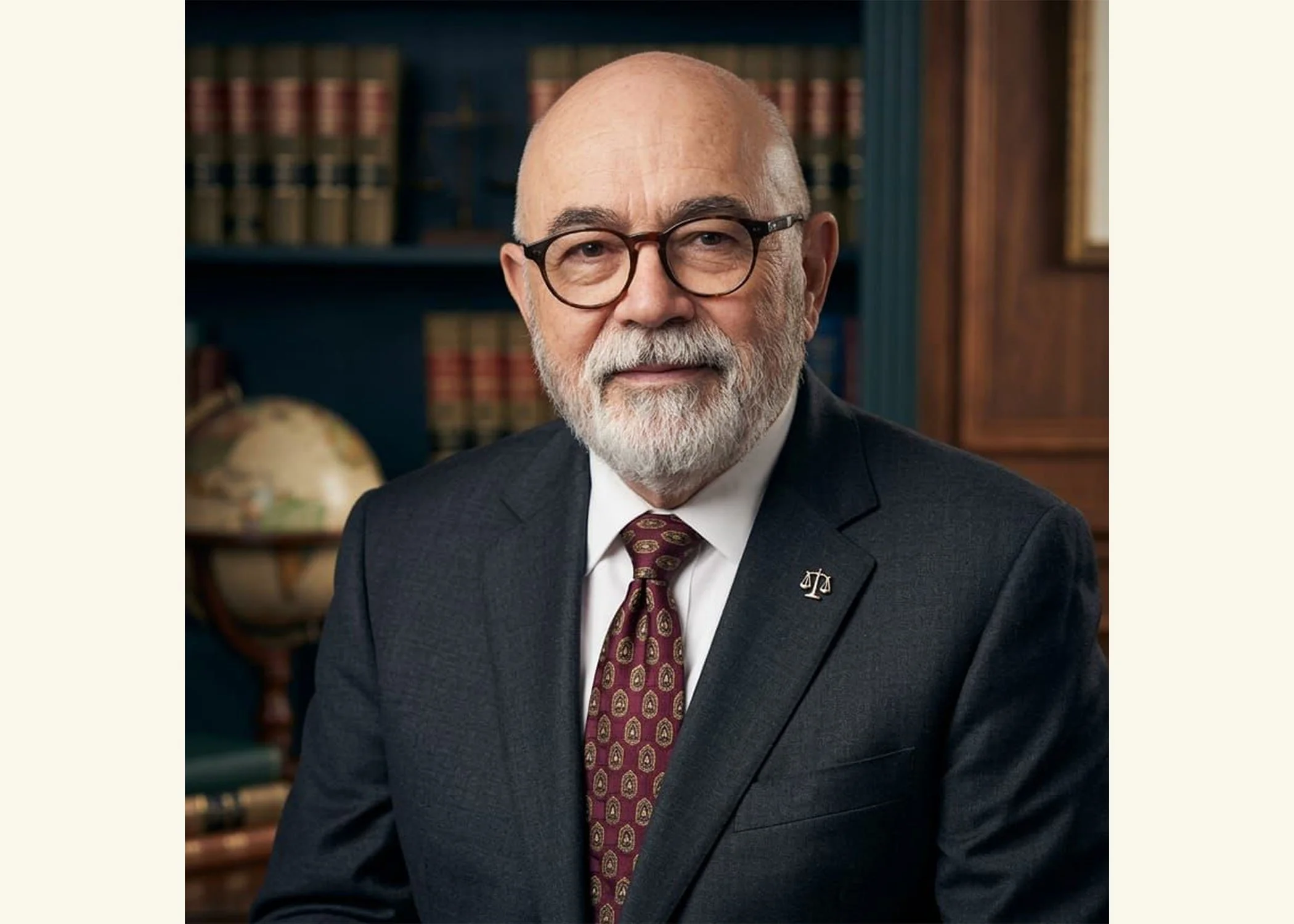 Lawyer Carlos Rivers in a dark suit, crimson tie, scales-of-justice pin, and glasses; he has a trim beard and is lightly smiling; in the background are a globe and an office with legal books.