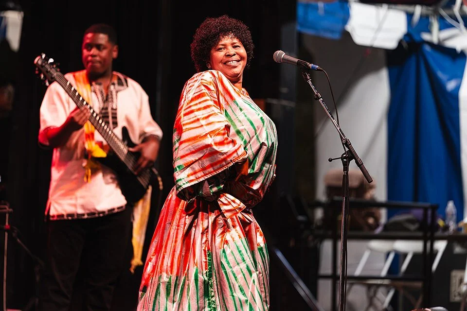 Sayon Camera singing at Richmond Folk Festival in a red and green patterned dress, smiling into the camera, with her bass player behind her.
