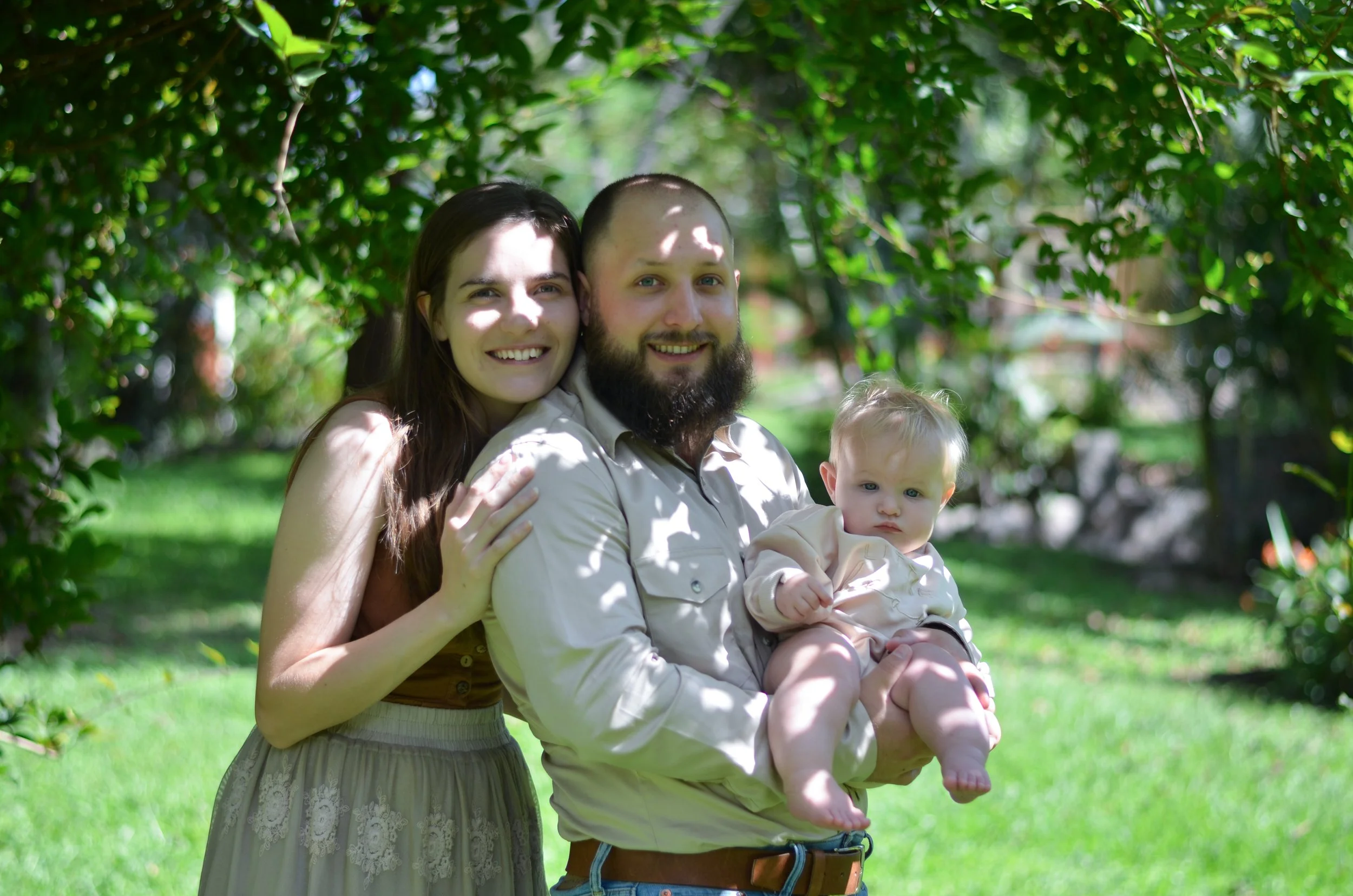 A family of three standing outdoors in a green, leafy park, smiling at the camera. The woman has long dark hair and is wearing a sleeveless top and skirt. The man has a beard, short hair, and is holding a young child with blonde hair, dressed in light-colored clothing.