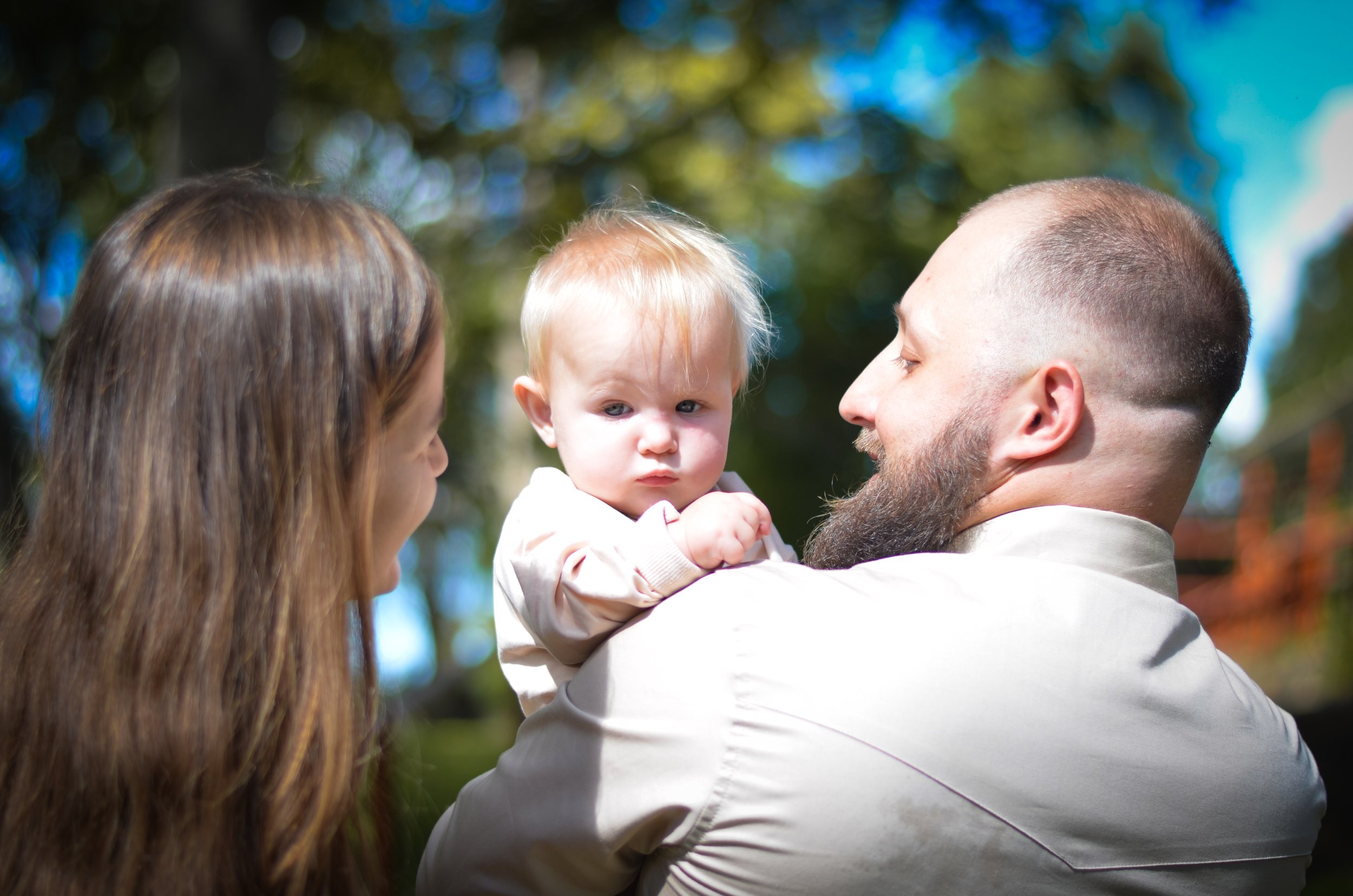 A man with a beard holds a baby girl, with a woman facing them, in an outdoor setting with trees and blue sky in the background.