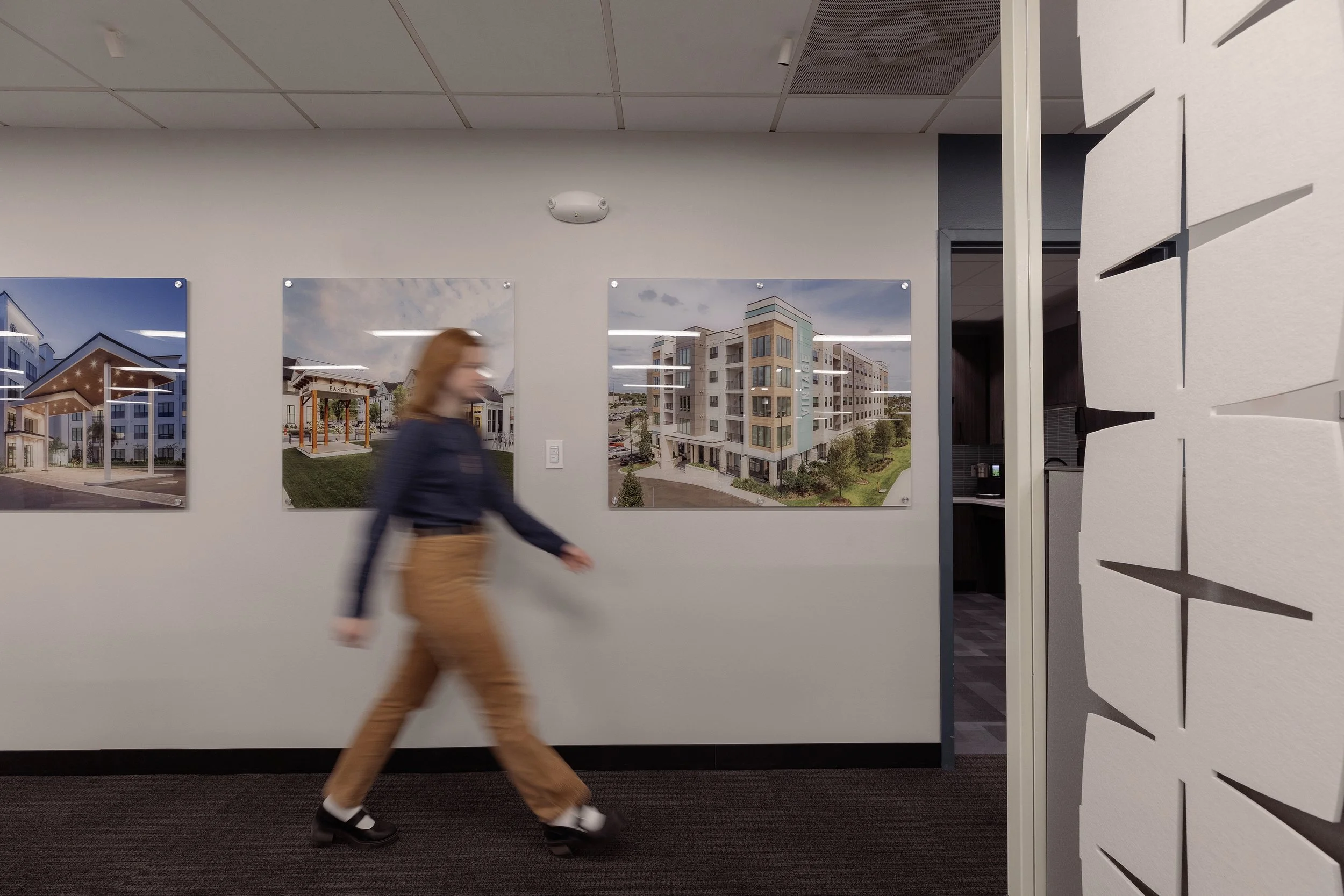 A woman walking past framed pictures of modern apartment buildings on a white wall in an indoor setting.
