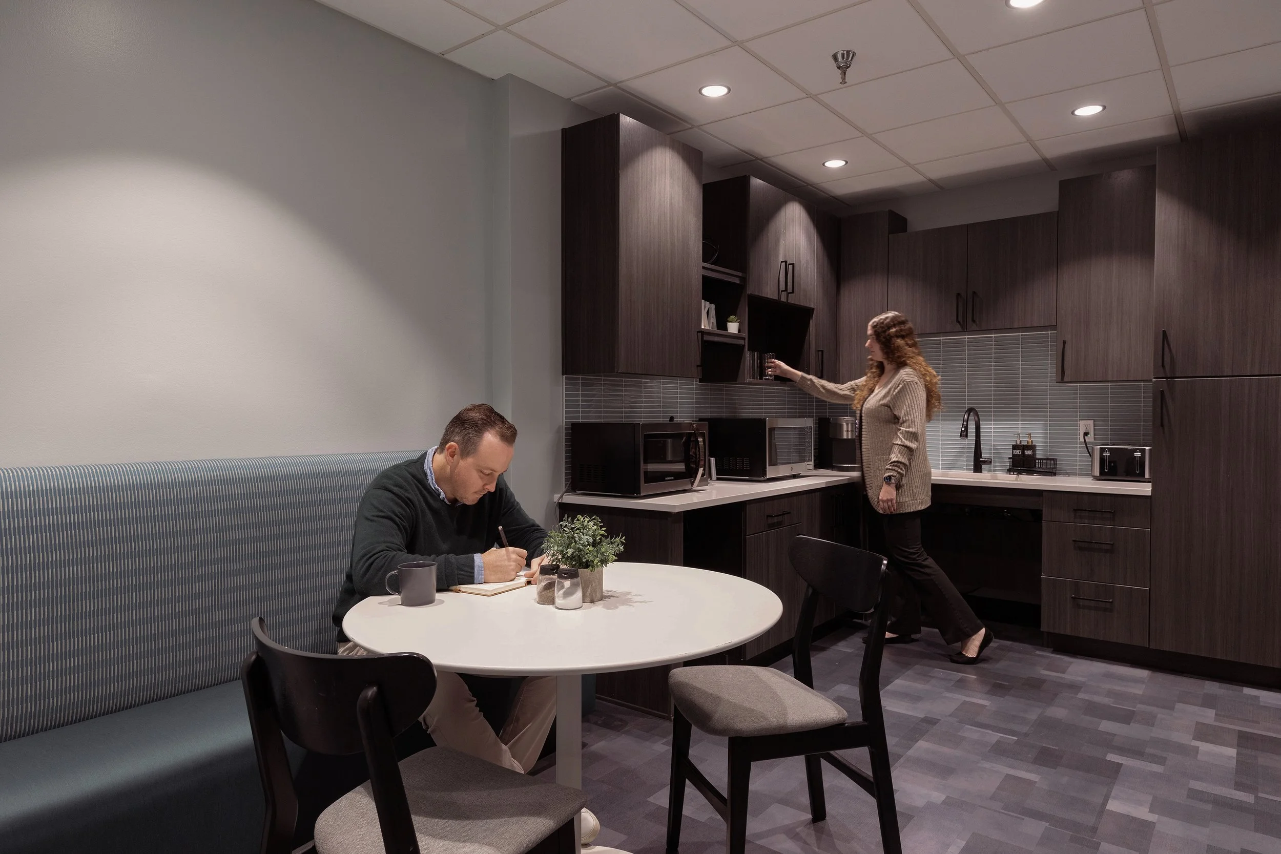 A man sitting at a table writing in a notebook with a mug beside him, and a woman standing near kitchen cabinets reaching for a glass, in a modern kitchen with dark wood cabinets and gray tile backsplash.