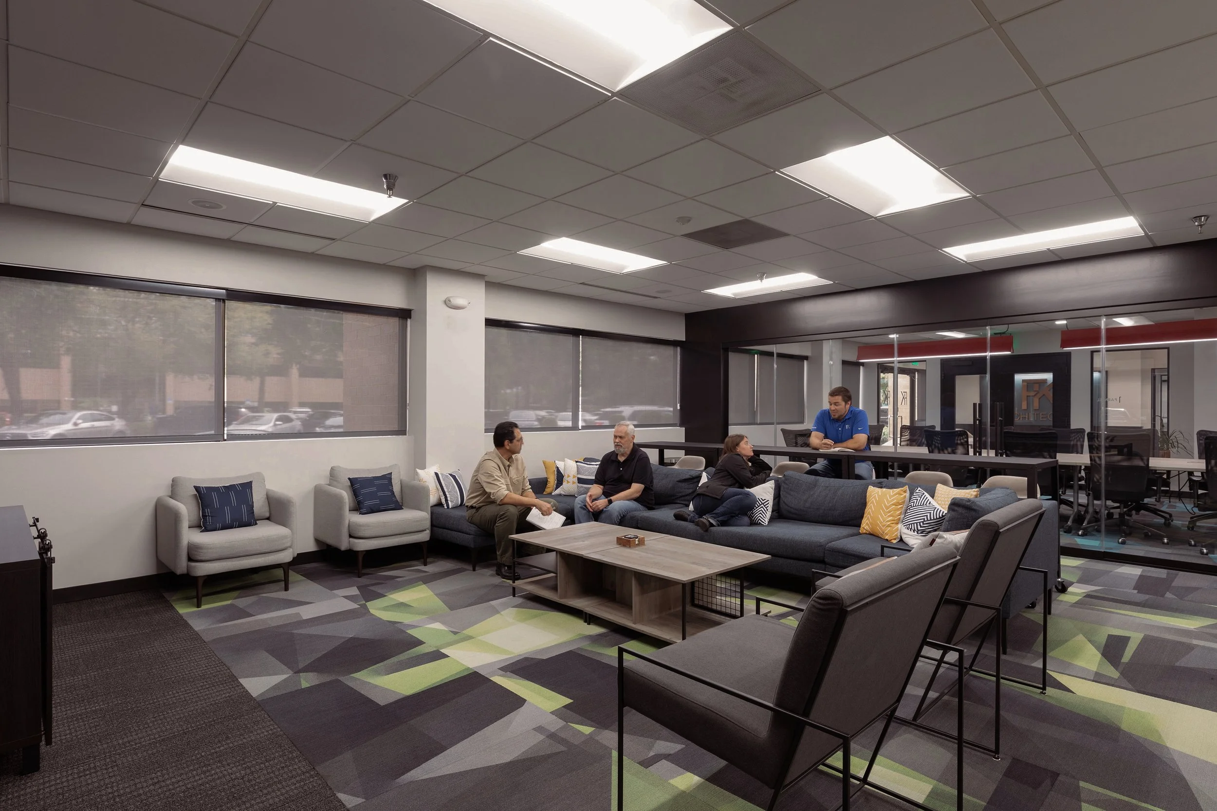 A waiting area in an office with modern gray sofas, colorful pillows, and a coffee table, with four people engaged in conversation and a man standing at a high table.