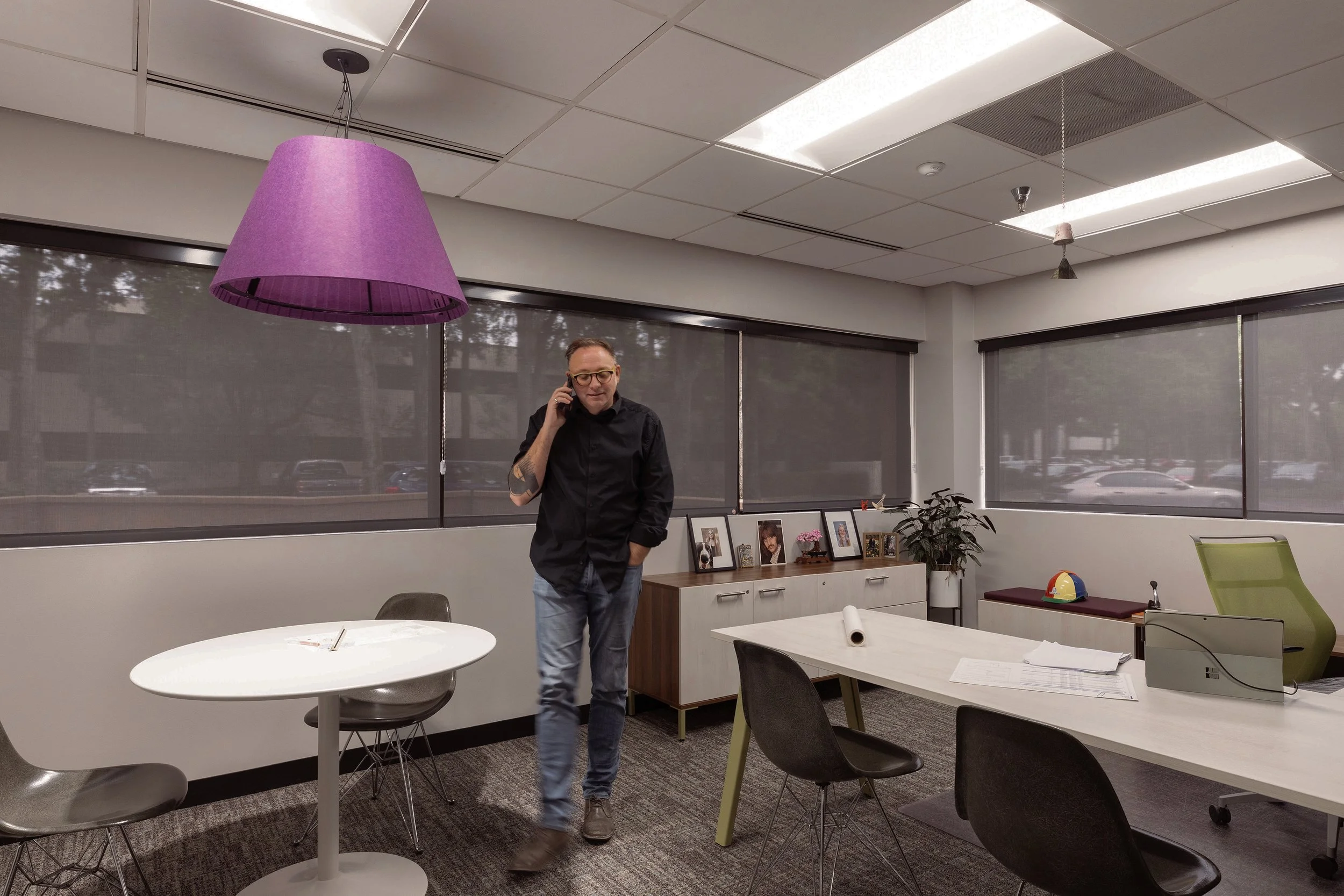 Man standing in an office with large windows, talking on his phone, surrounded by tables, chairs, and office decor.