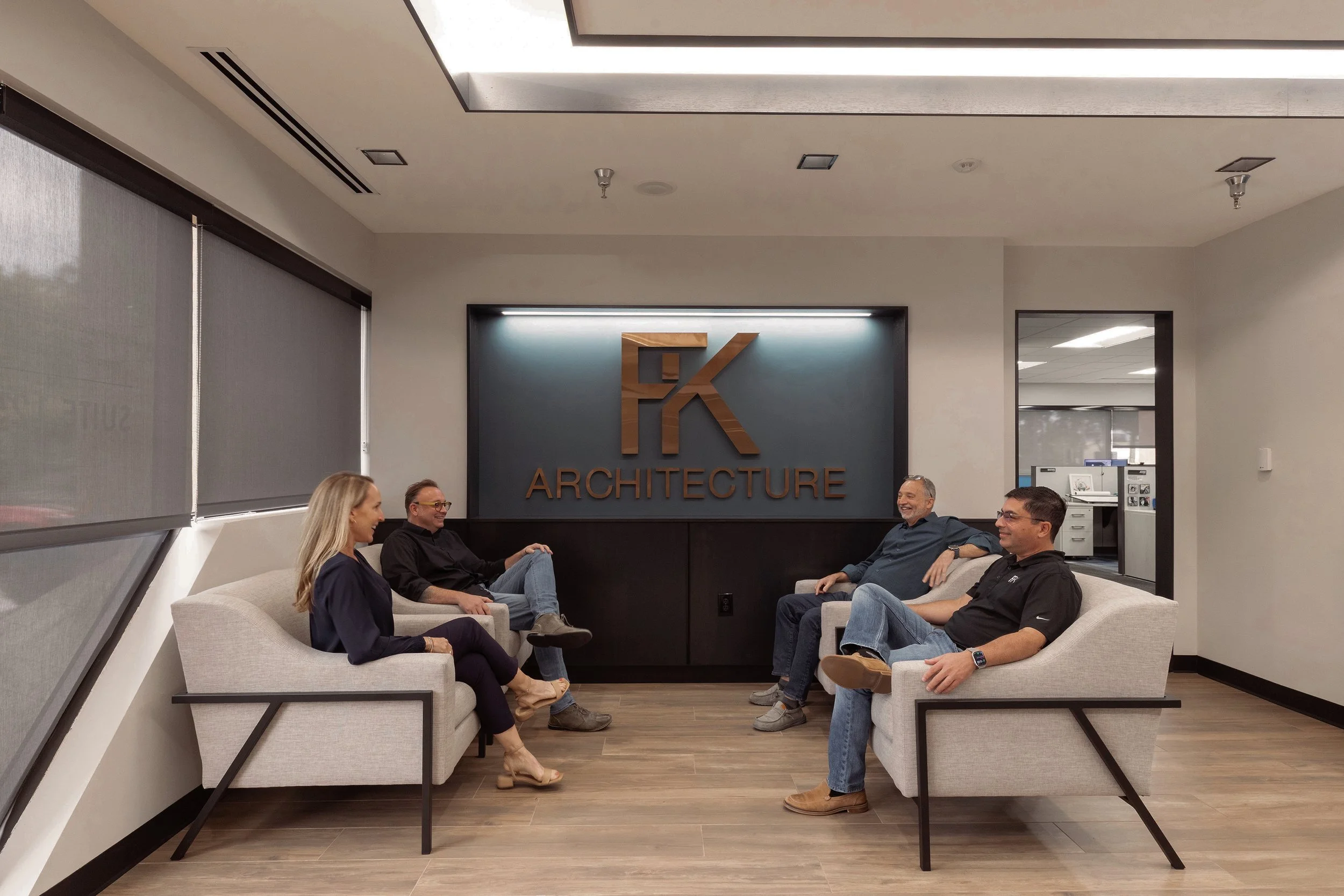 Five people sitting on sofas in a modern office waiting area, engaged in conversation, with a large sign reading 'FK Architecture' on the wall behind them.