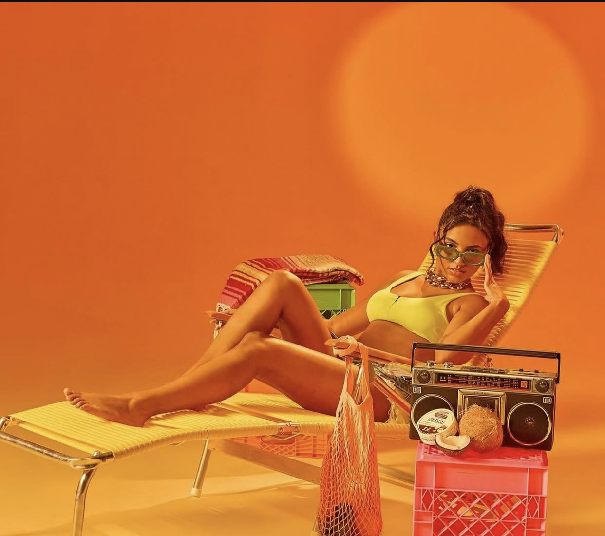 A woman lounging on a yellow sunbed with an orange background, wearing sunglasses and a yellow top, with a vintage boombox, coconut, and other beach items nearby.