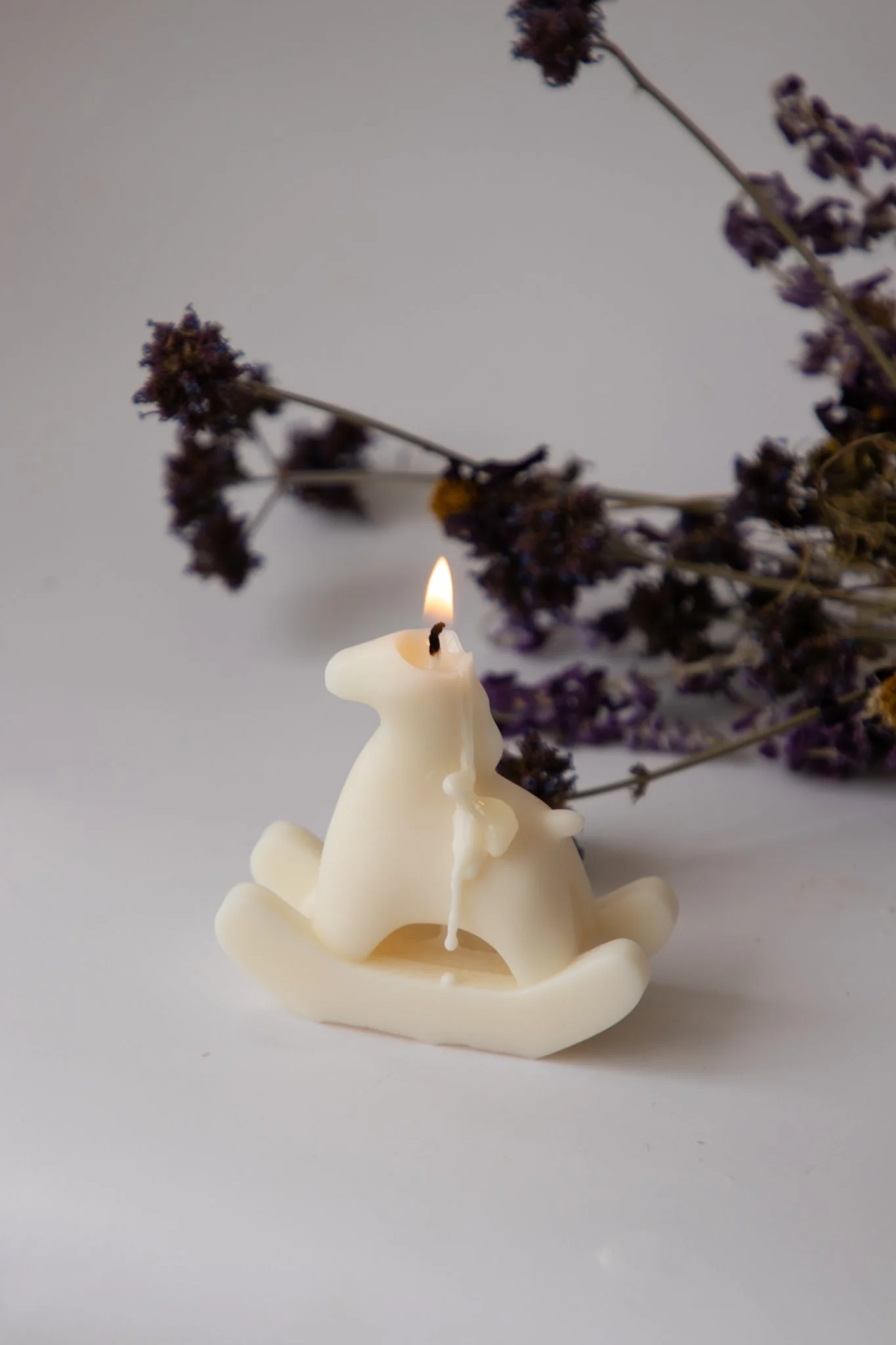 Close-up of handmade rocking horse candle burning, on white background with dried florals