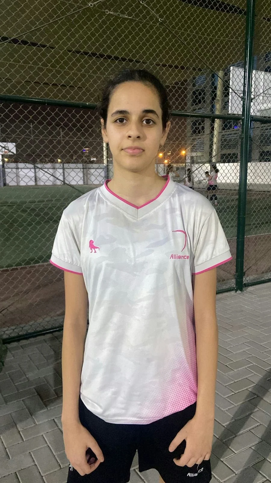 A young woman wearing a white sports T-shirt with pink accents and black shorts, standing in front of a chain-link fence on a sports field at night.