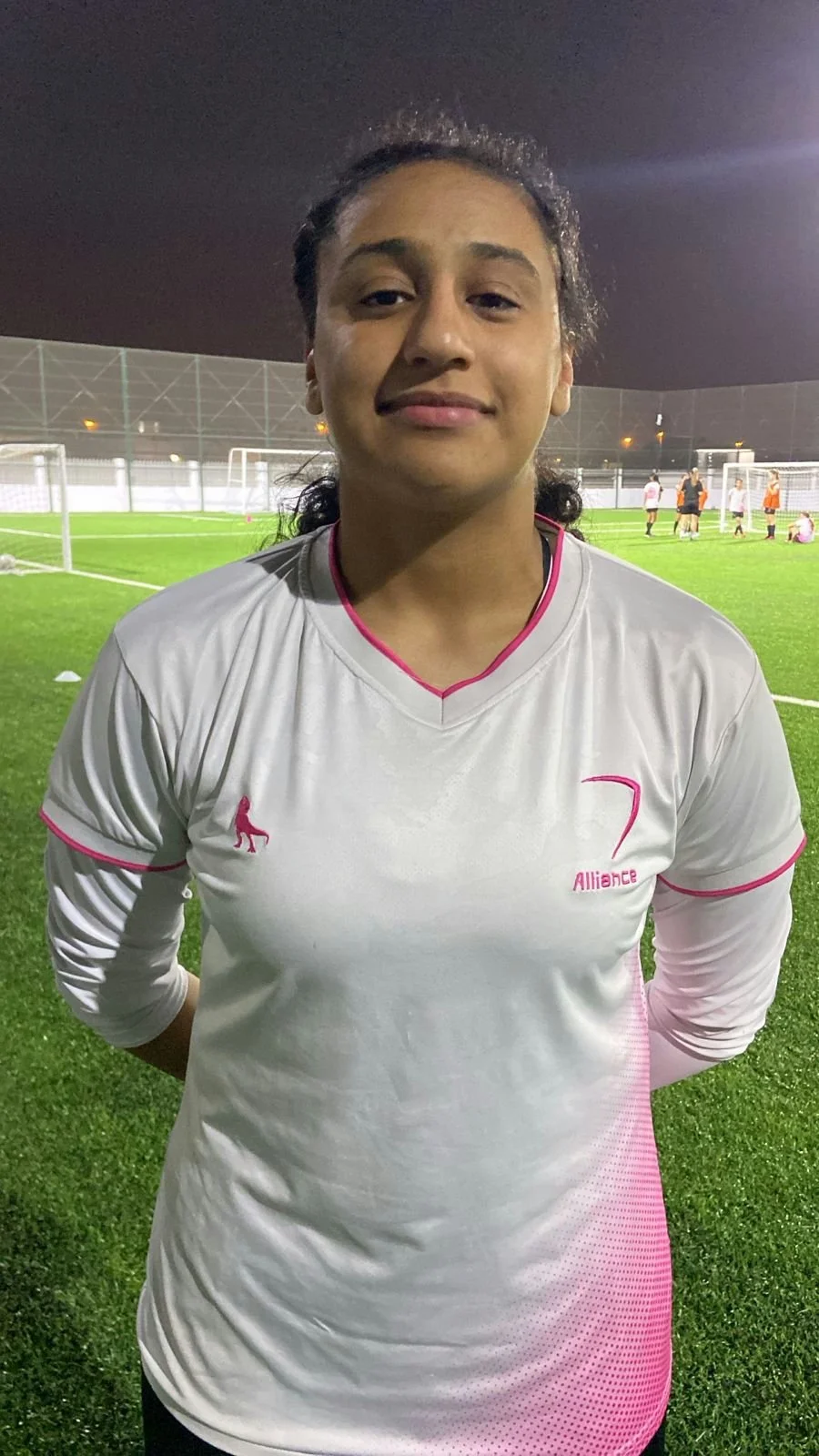 A young woman with dark hair tied back, wearing a white and pink soccer jersey, standing on a green soccer field at night with a confident expression.