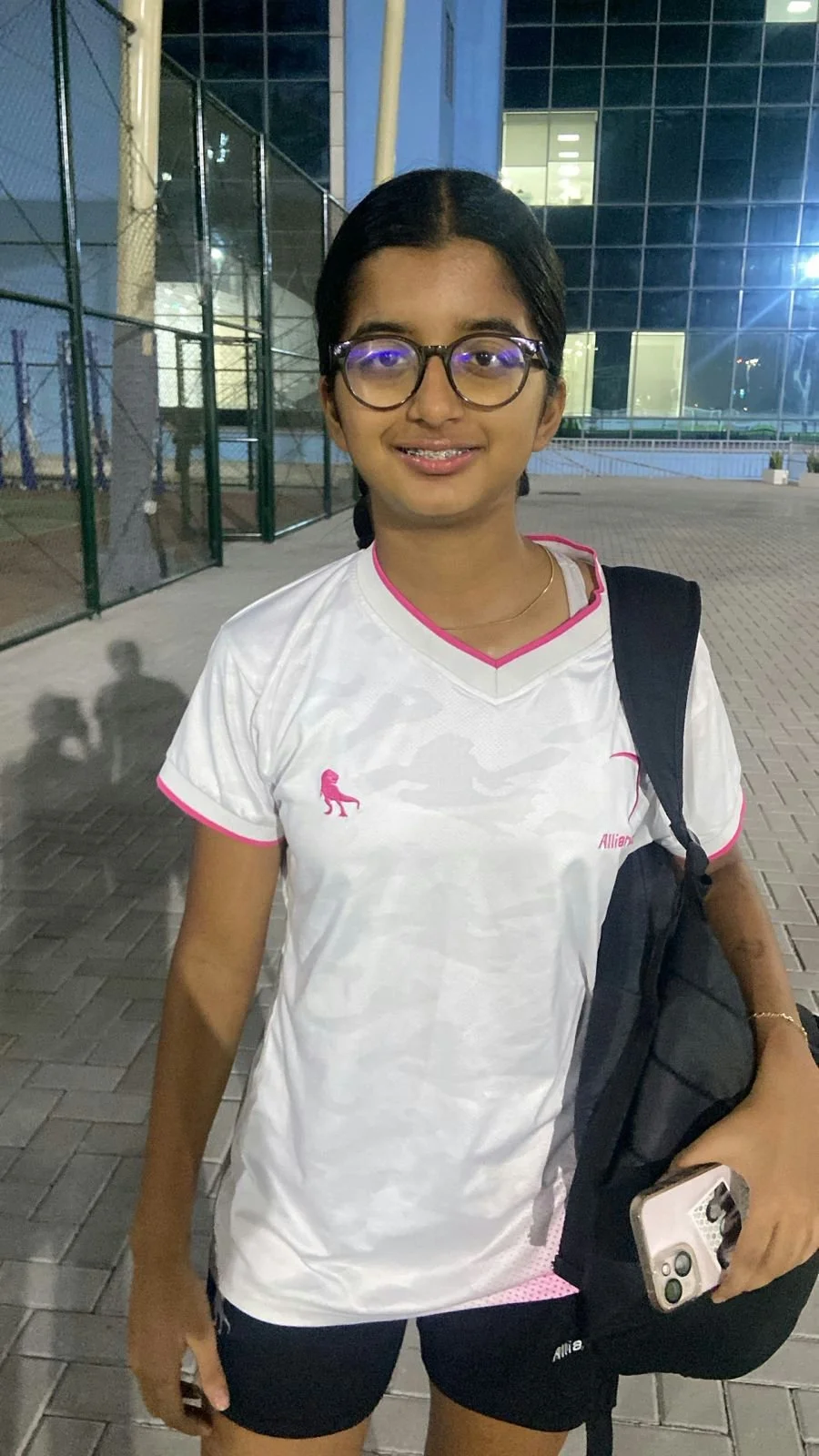A young girl with glasses and braces standing outdoors at night, wearing a white sports jersey with pink accents and a small pink lion logo, carrying a black backpack and holding a smartphone.