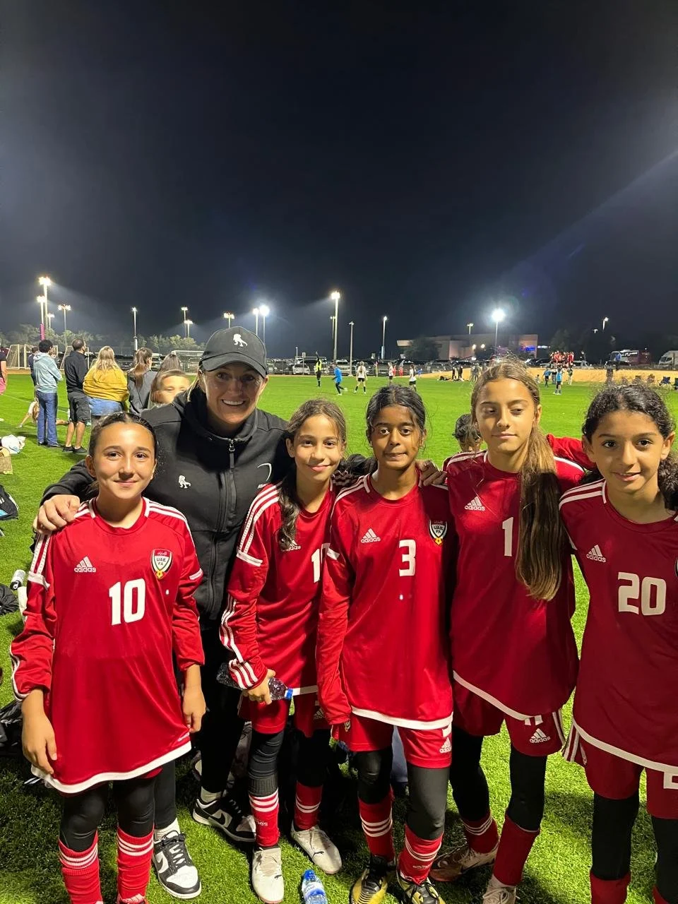 Young female soccer players in red uniforms with a coach, standing on a soccer field at night after a game with lights and other players in the background.