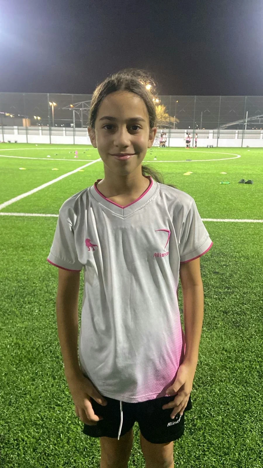 A young girl standing on a soccer field at night, wearing a white sports jersey with pink accents and black shorts, smiling.