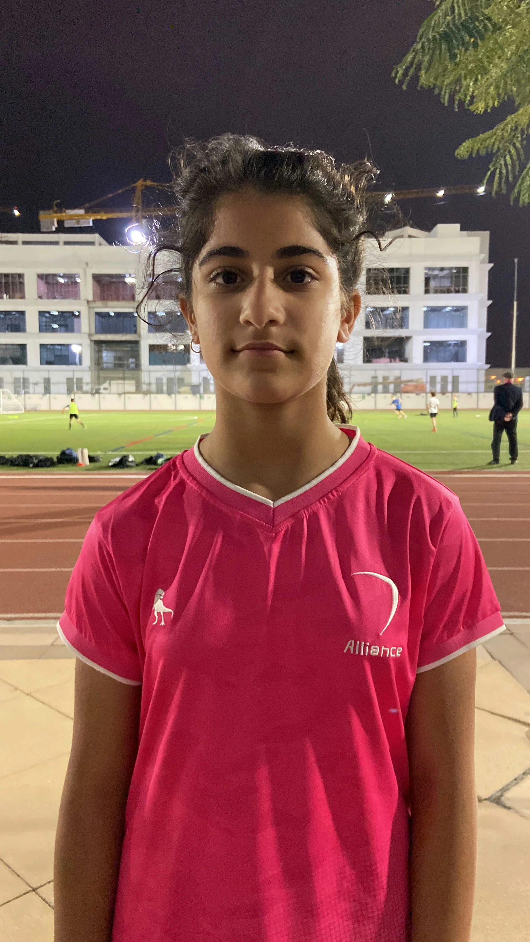 Young girl in pink sports jersey standing on track at night with a football field and building in the background.