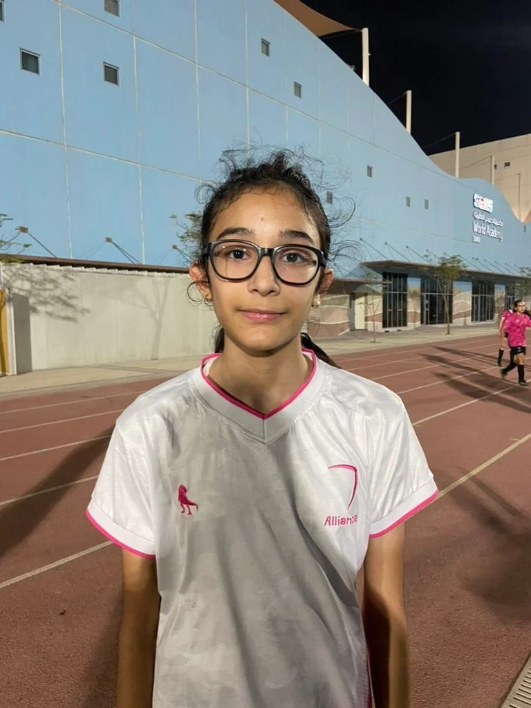 A young girl with glasses and long dark hair tied back, wearing a white sports jersey with pink accents, standing on a running track at night.