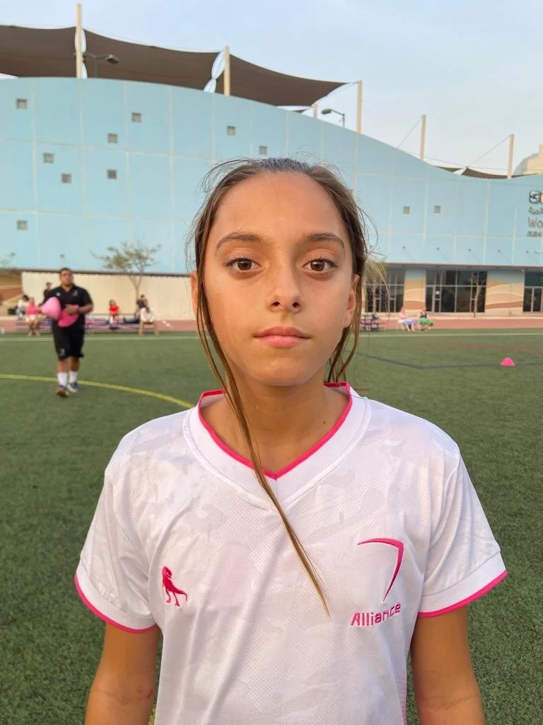 A girl in a white sports jersey standing on a soccer field with a large building, other children, and a man in the background.
