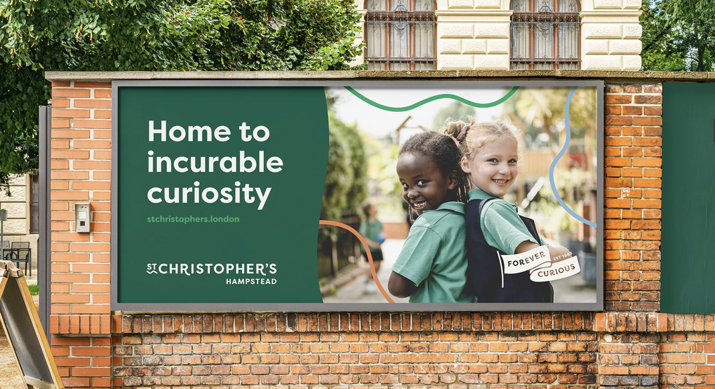 Outdoor billboard campaign for St Christopher’s Hampstead featuring two smiling children in school uniform alongside bold typography promoting curiosity, displayed on a brick wall in a leafy urban setting.