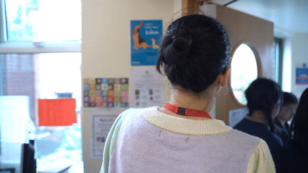 Back view of a woman with dark hair in a bun wearing a light-colored sweater and a red lanyard, standing in a room with others near a door with an oval window.