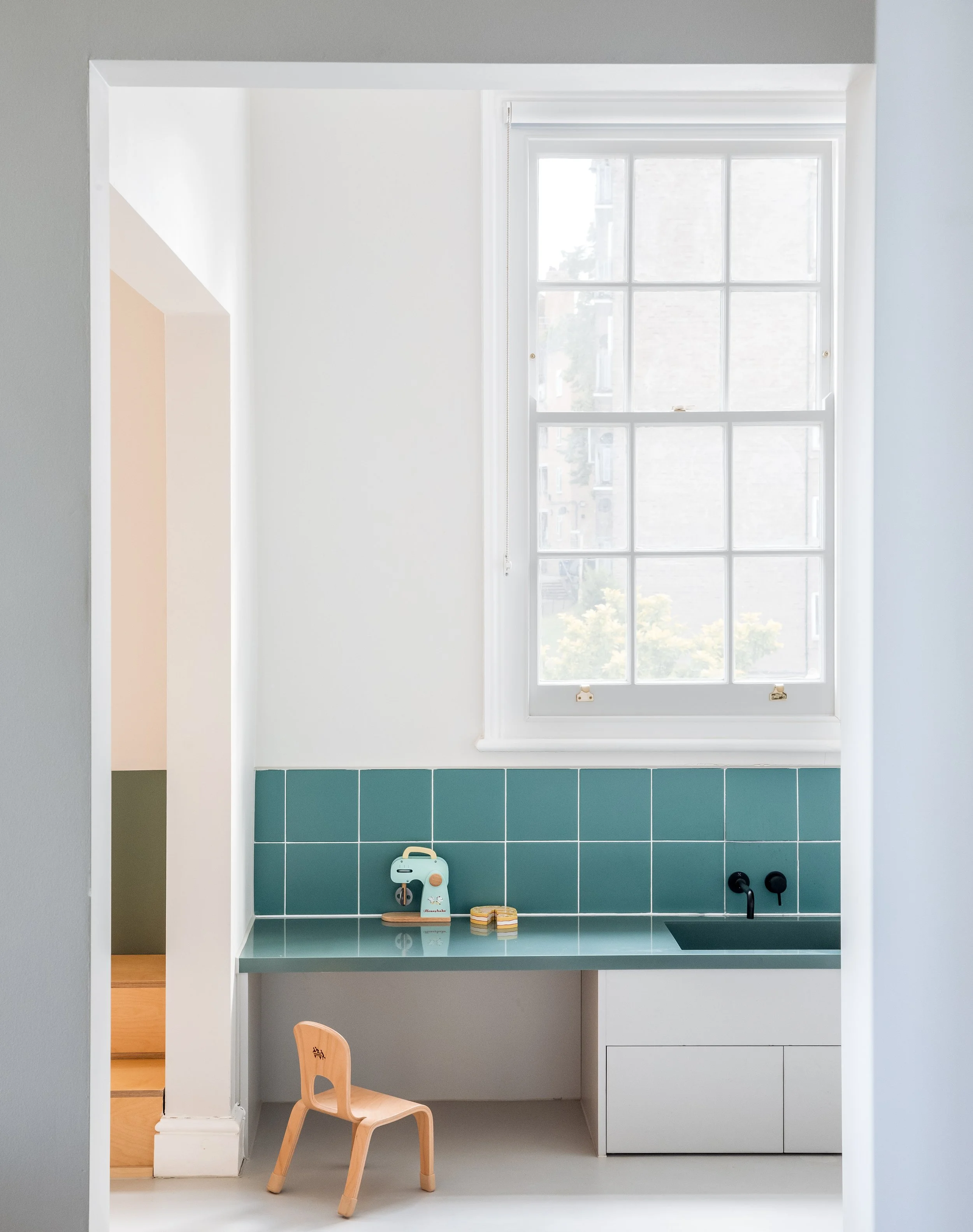 Minimal child-sized learning kitchen with pastel blue tiles, timber chair, and integrated play features designed to support independence and early years learning within a calm interior environment.