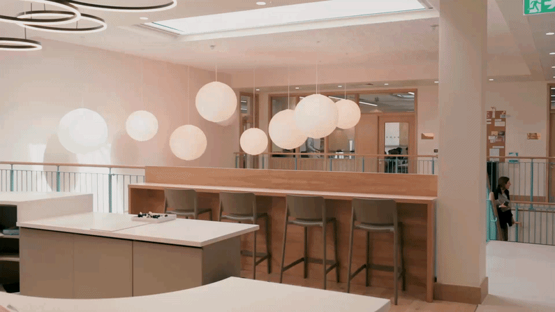 Modern interior of a coworking or lounge area with hanging spherical pendant lights, high bar seating, and a person walking in the background.