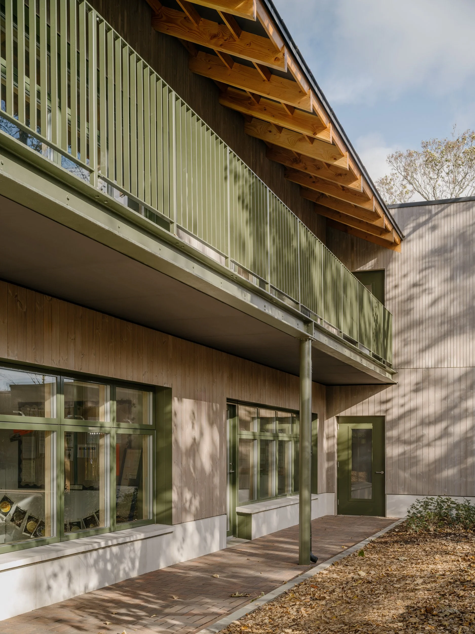 Contemporary timber-clad school building with green metal balcony, exposed roof structure, and shaded external circulation spaces designed for sustainable educational architecture.