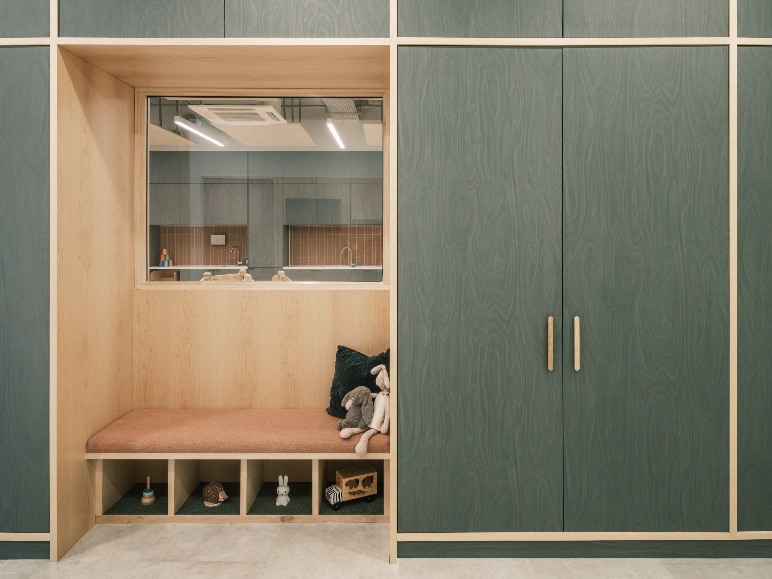 Custom nursery storage wall featuring deep green timber cabinetry, an integrated upholstered reading nook, open toy cubbies, and natural wood detailing designed to support organised, child-friendly early years learning environments.