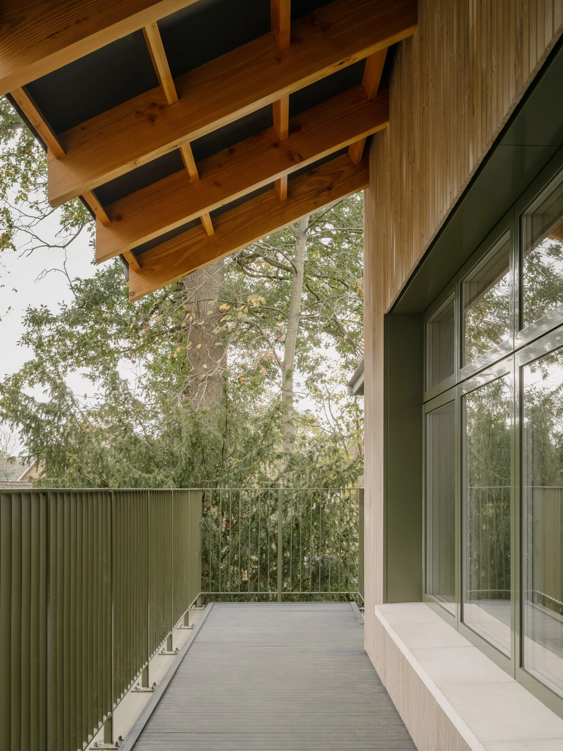 External school balcony with timber roof structure, green metal balustrade, and views into surrounding trees, designed as a sheltered outdoor learning and circulation space.