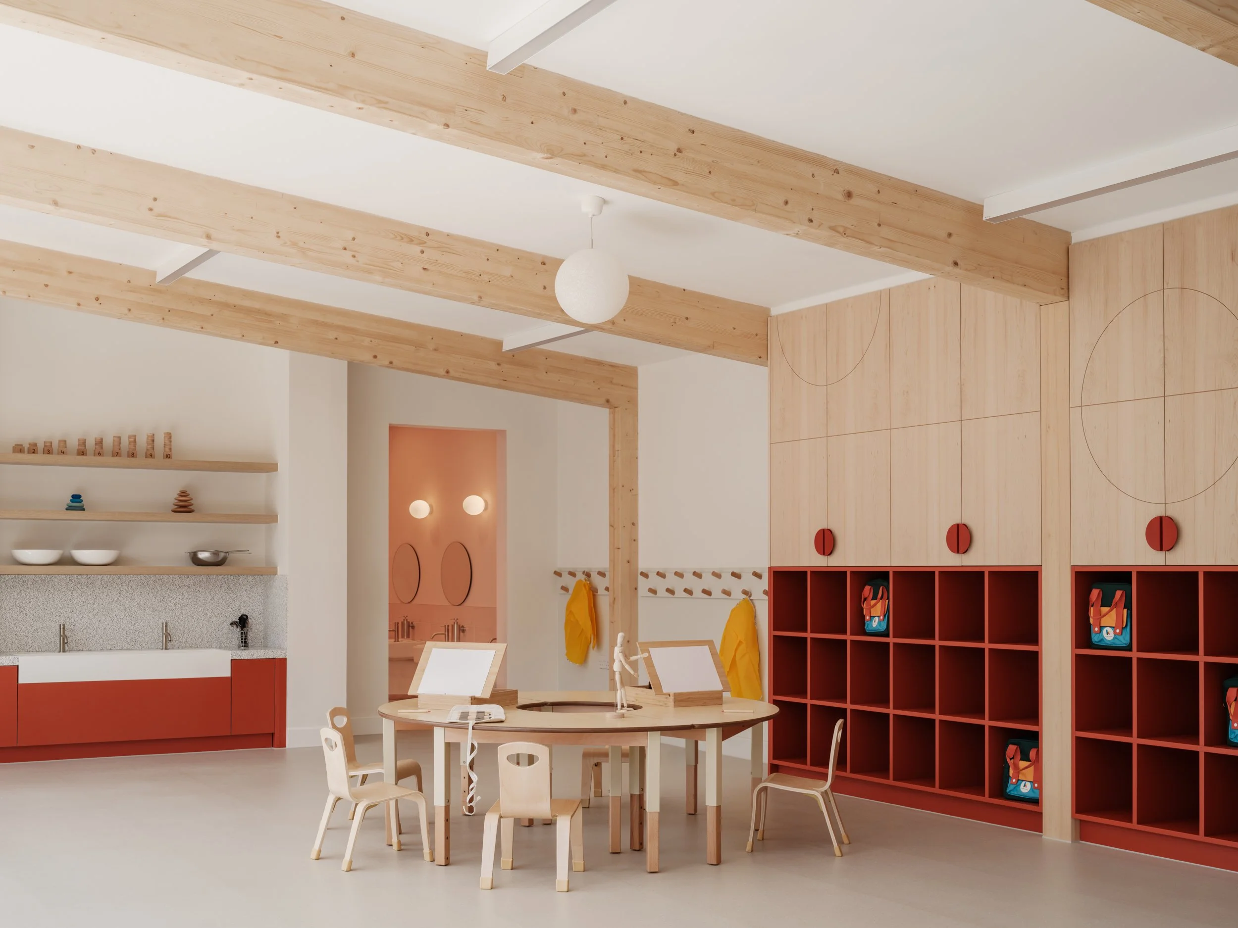Minimal early years classroom featuring integrated timber and red joinery, child-scale storage, soft play elements, and circular openings designed to support independent learning and imaginative play.