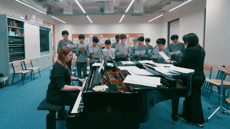 A music class with a woman playing grand piano while a group of children sing and follow a music teacher in a rehearsal room.