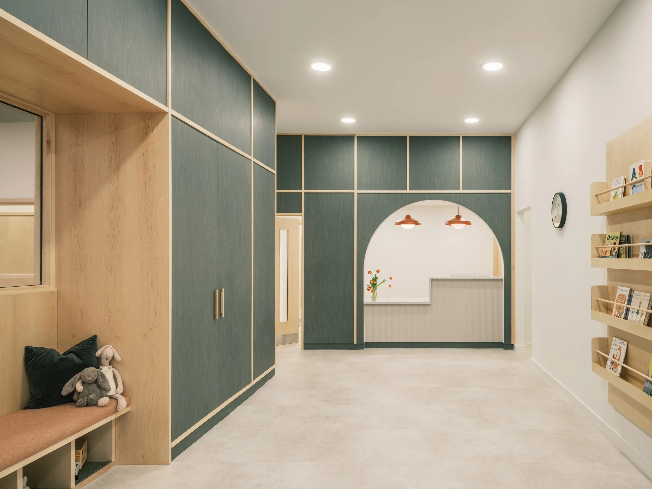 Contemporary nursery reception and circulation space featuring integrated timber seating, deep green cabinetry, an arched reception opening, children's book displays, and warm natural materials designed to create a welcoming early years environment.