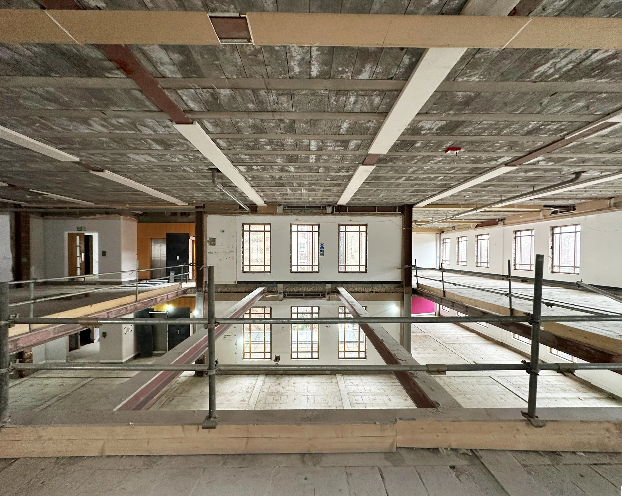 Interior of a building under construction with exposed wooden beams, gray and white flooring, and multiple large windows.